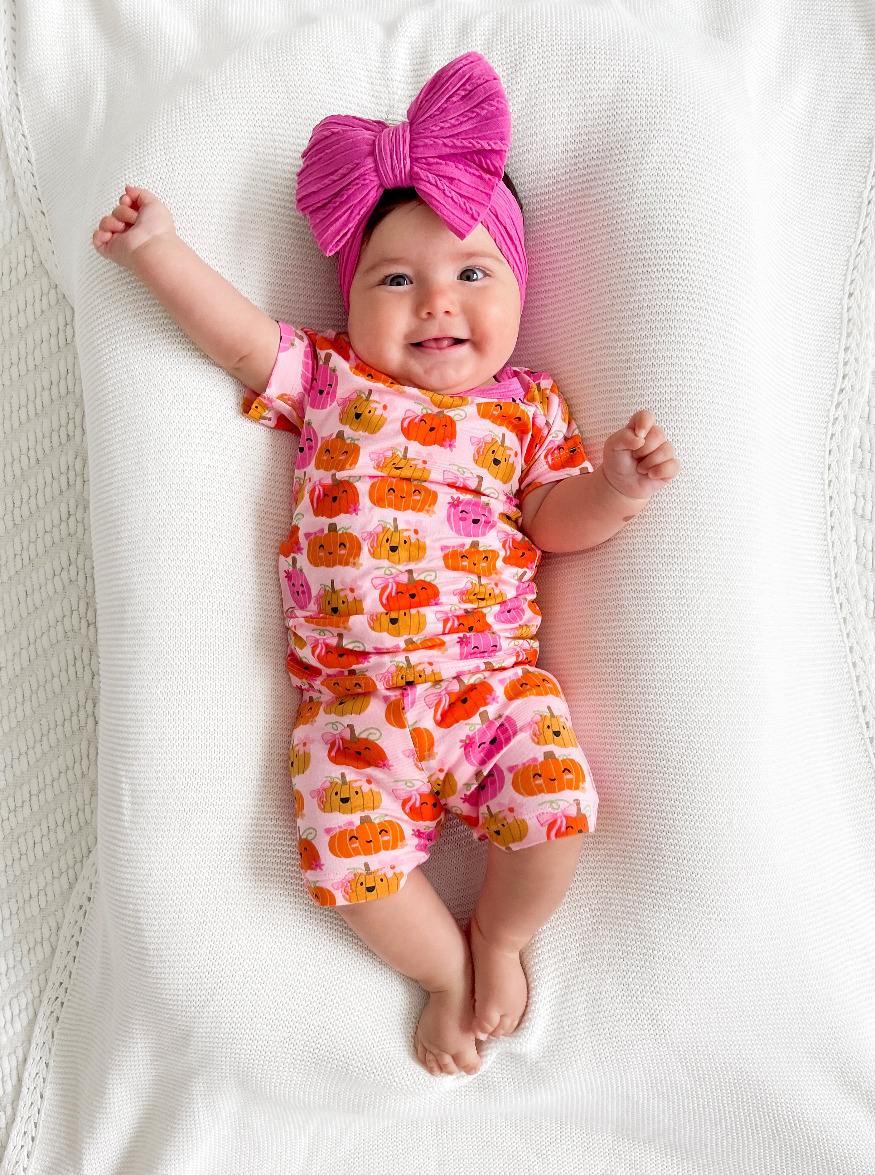 Smiling baby in a pumpkin-patterned outfit with a large pink bow, lying on a soft white blanket.