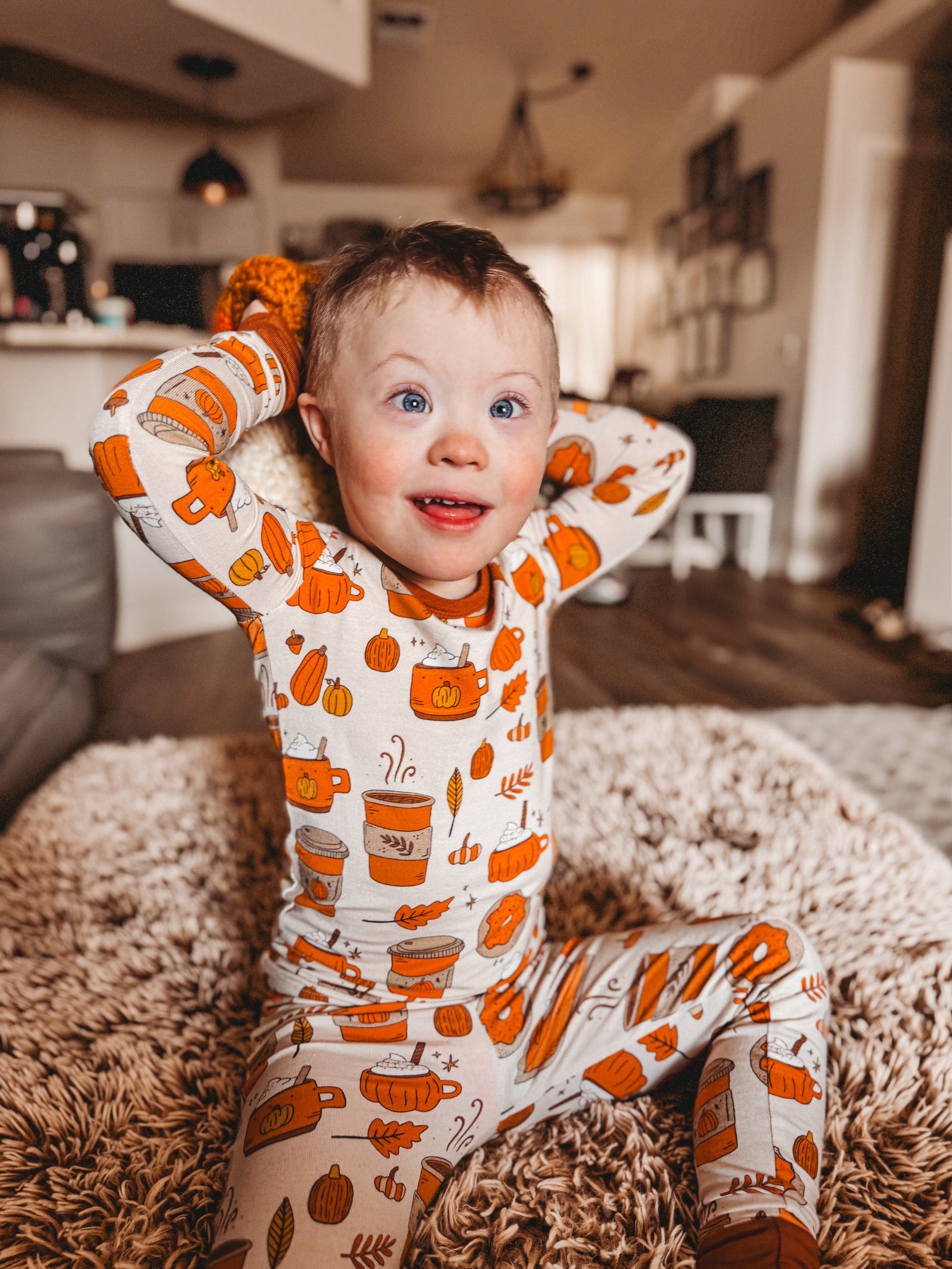 Child in festive pajamas with pumpkins, sitting on a rug, smiling joyfully in a cozy home setting.