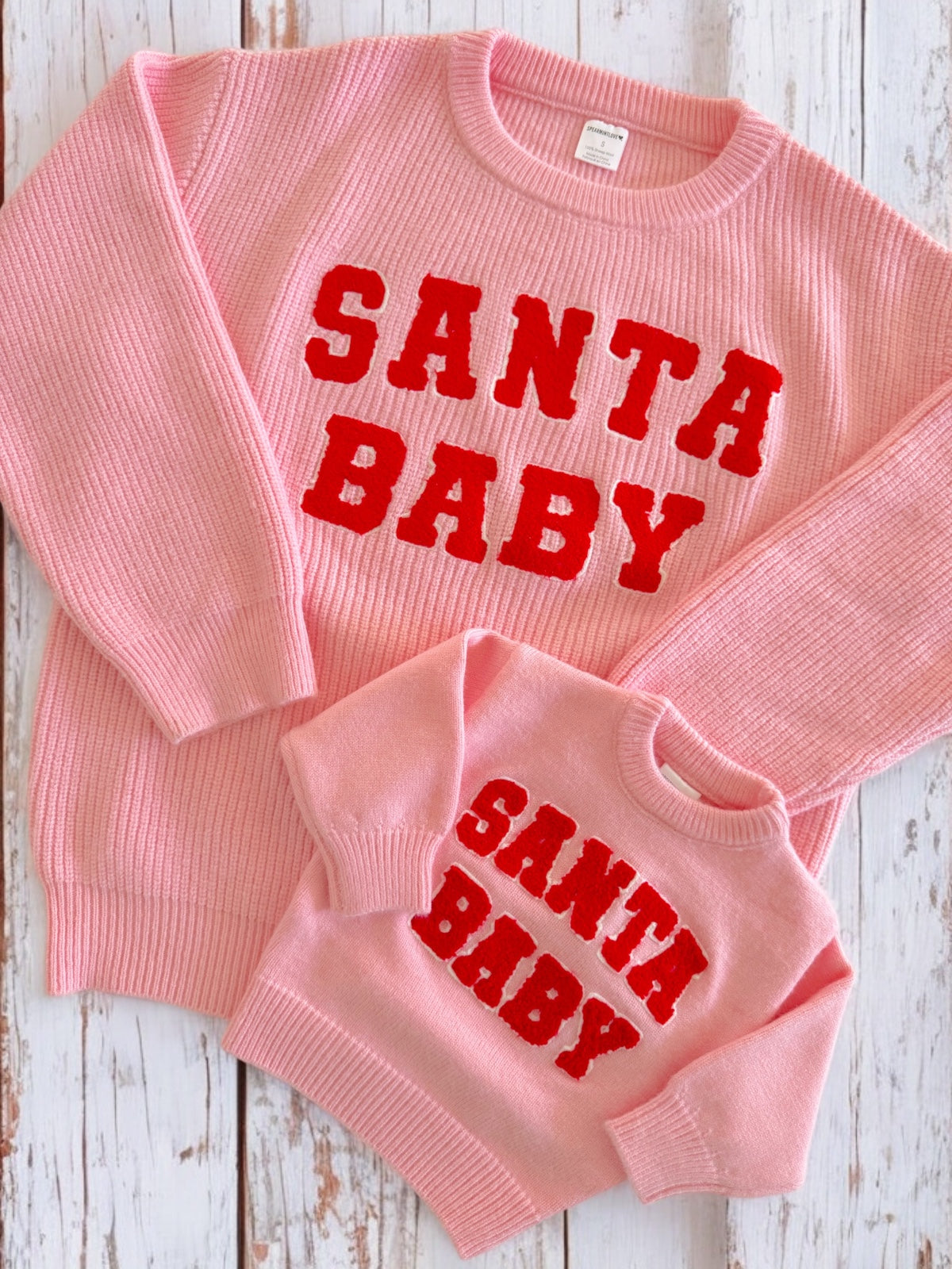 Matching pink sweaters with "SANTA BABY" in red text for adults and children, displayed on a wooden background.