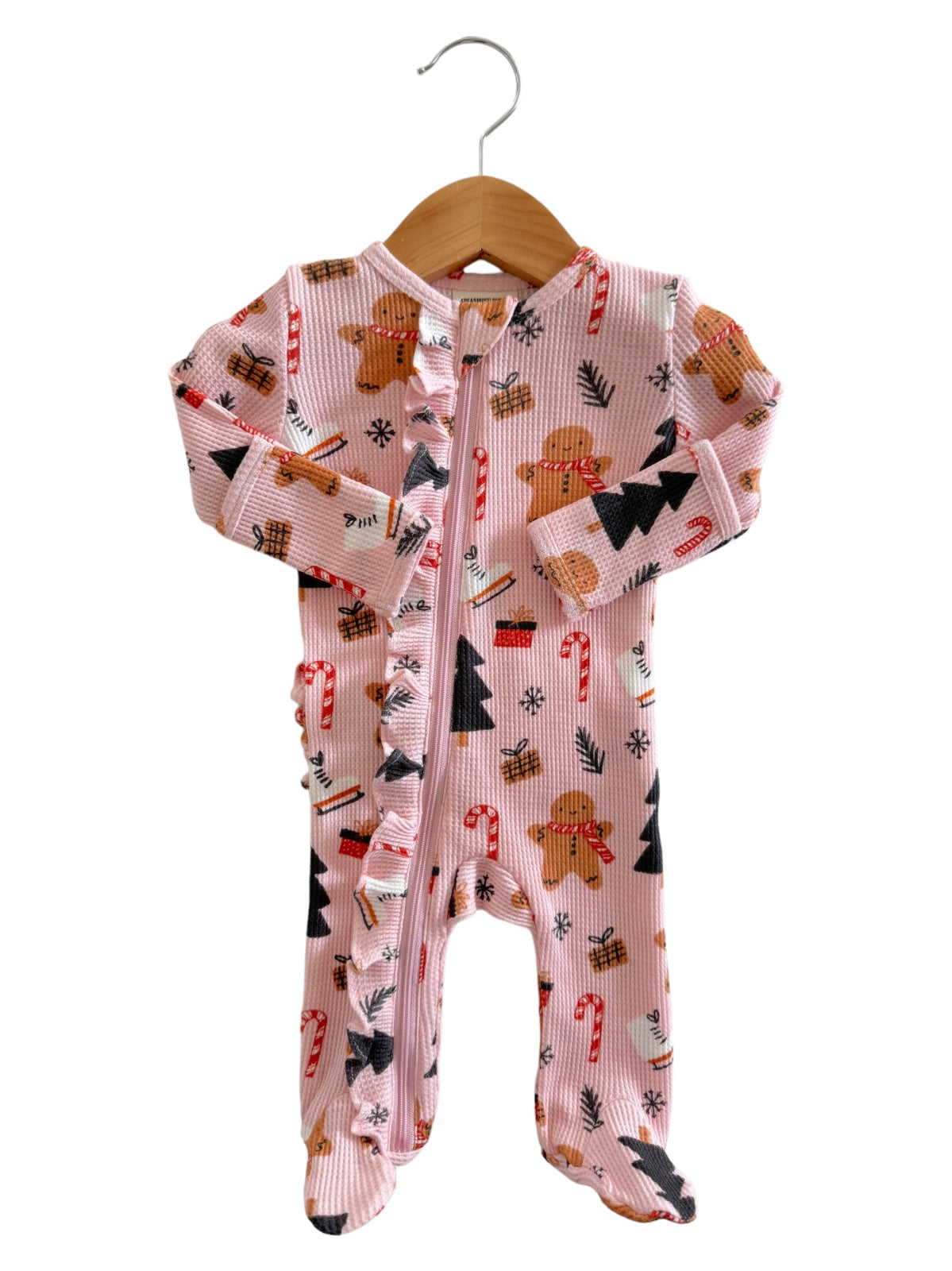 Pink holiday-themed pajama onesie with gingerbread, candy canes, and Christmas trees, hanging on a wooden hanger.