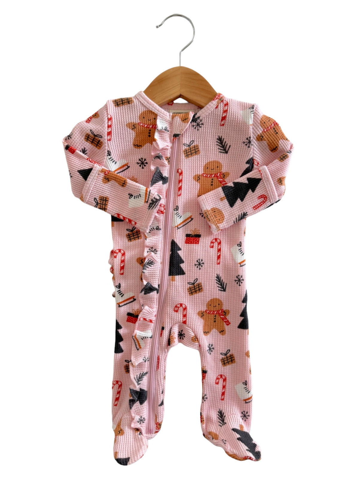 Pink holiday-themed pajama onesie with gingerbread, candy canes, and Christmas trees, hanging on a wooden hanger.