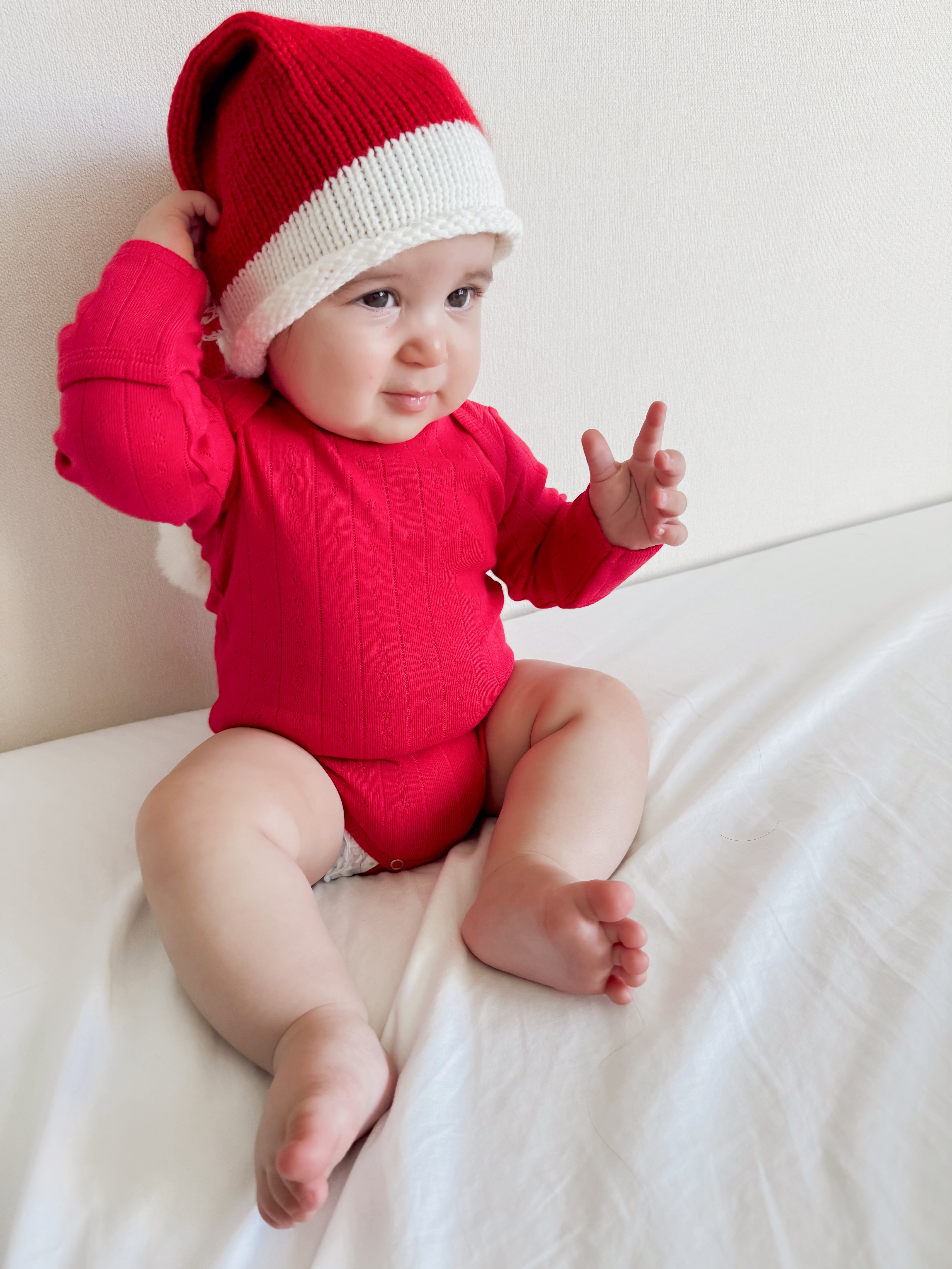 Baby in a red outfit and white hat, sitting on a white bed, smiling and playing with one hand raised.