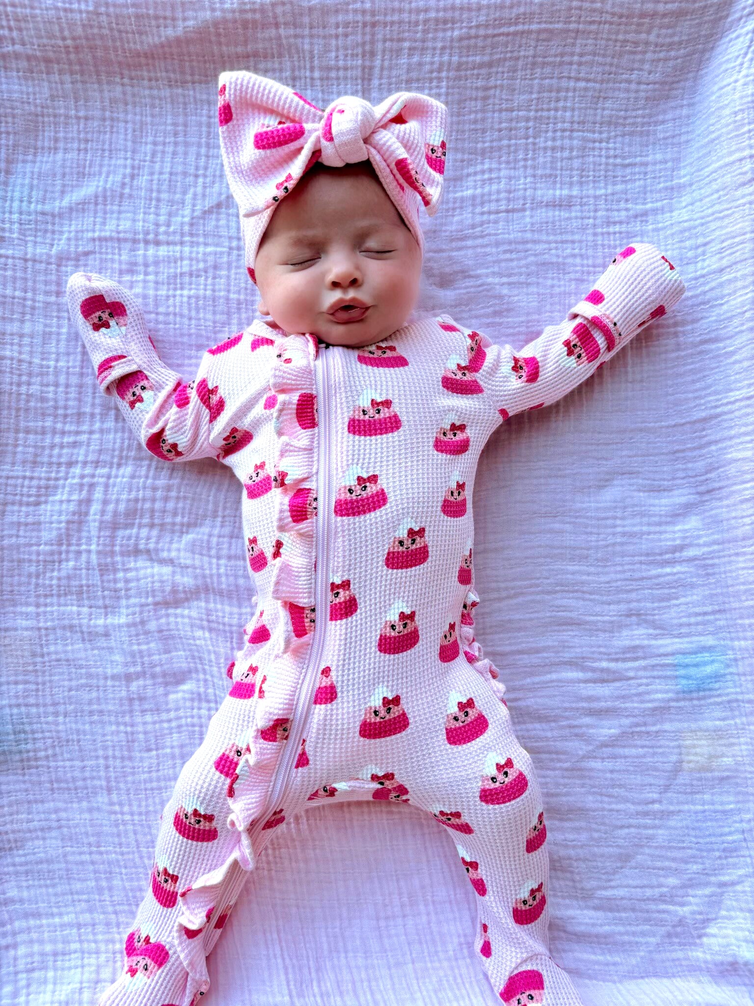 Baby in a pink cake-patterned onesie and bow, lying on a white fabric background with arms outstretched.