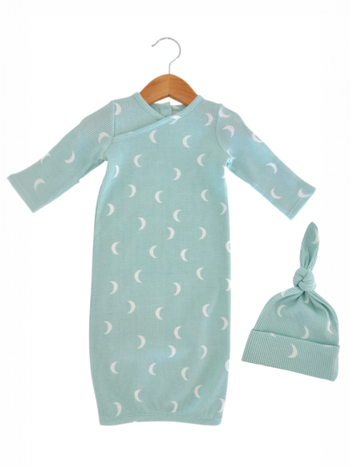 Mint green baby gown with white crescent moon pattern and matching knotted hat, displayed on a wooden hanger.