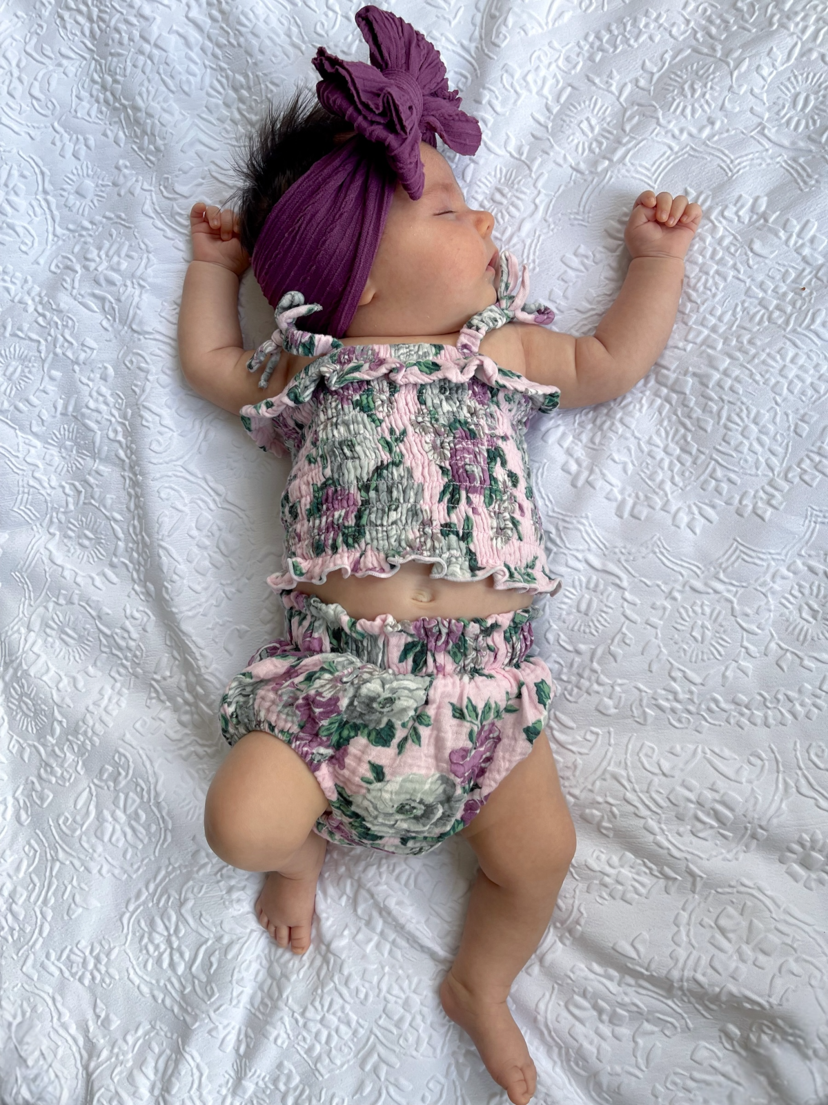 Baby girl dressed in floral outfit with a purple headband, lying on a textured white blanket.