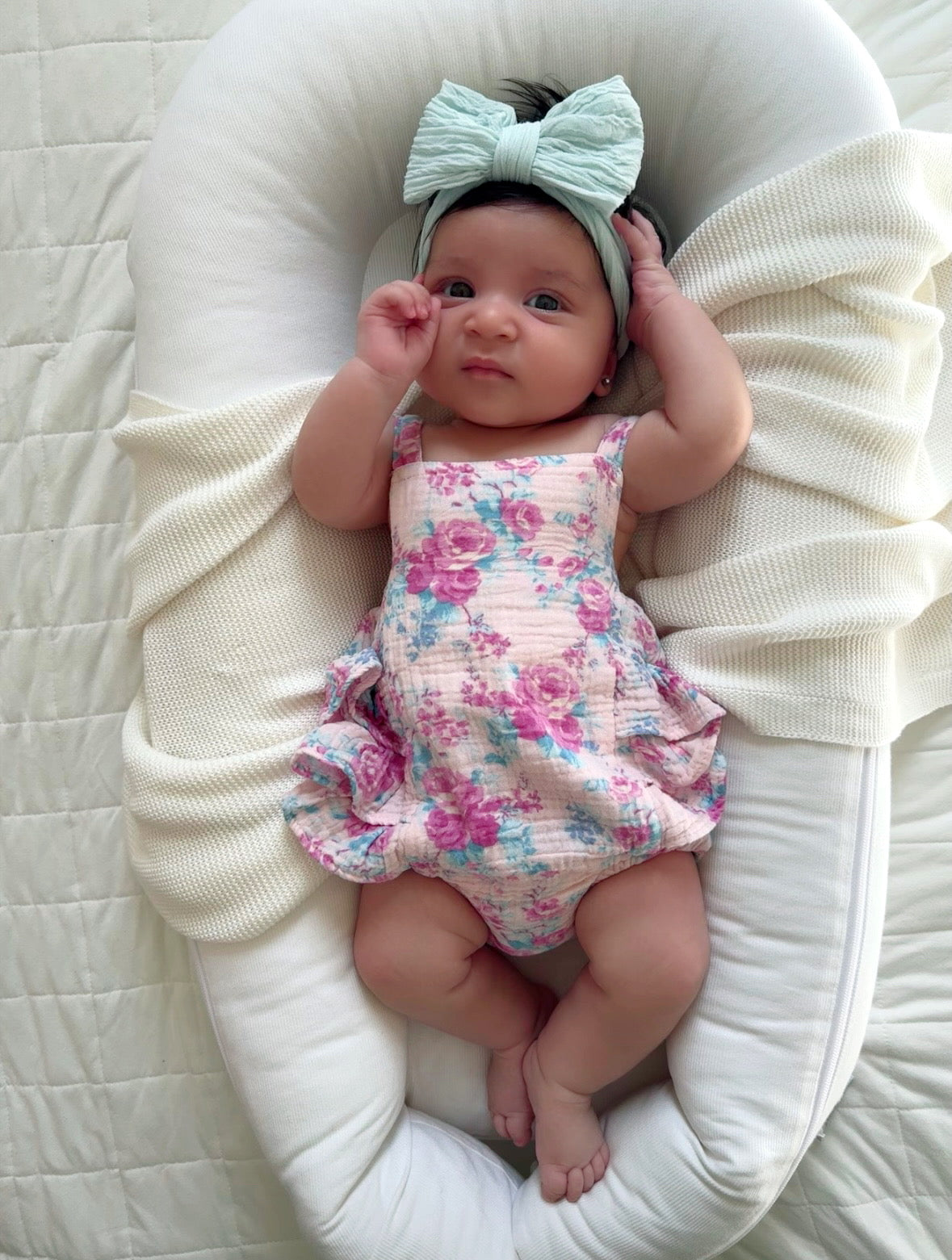 Baby girl in floral outfit and bow, resting peacefully on a cozy blanket. Bright, curious expression.
