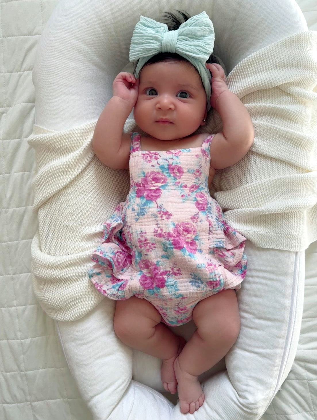 Baby girl in a floral romper with a mint bow, lying on a cozy blanket, looking curiously at the camera.