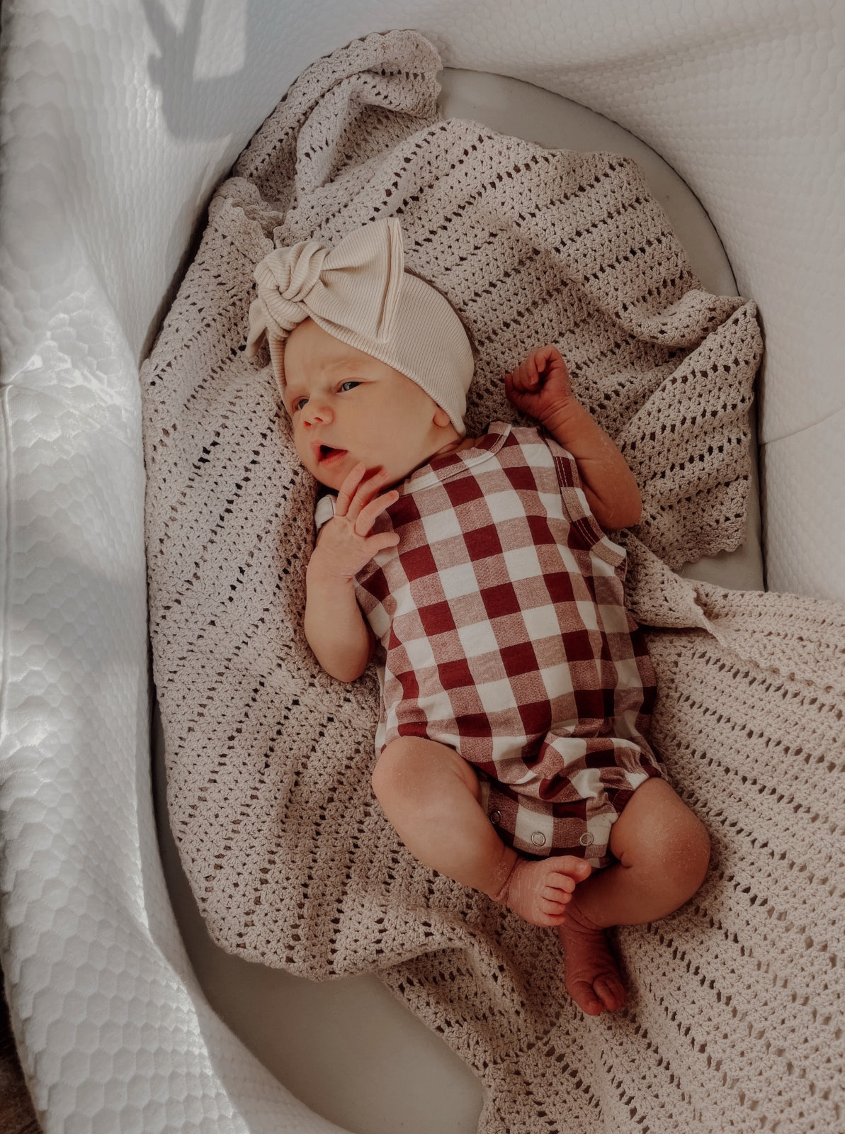Infant lying on a textured blanket, wearing a checkered romper and a bow headband. Soft, warm lighting.