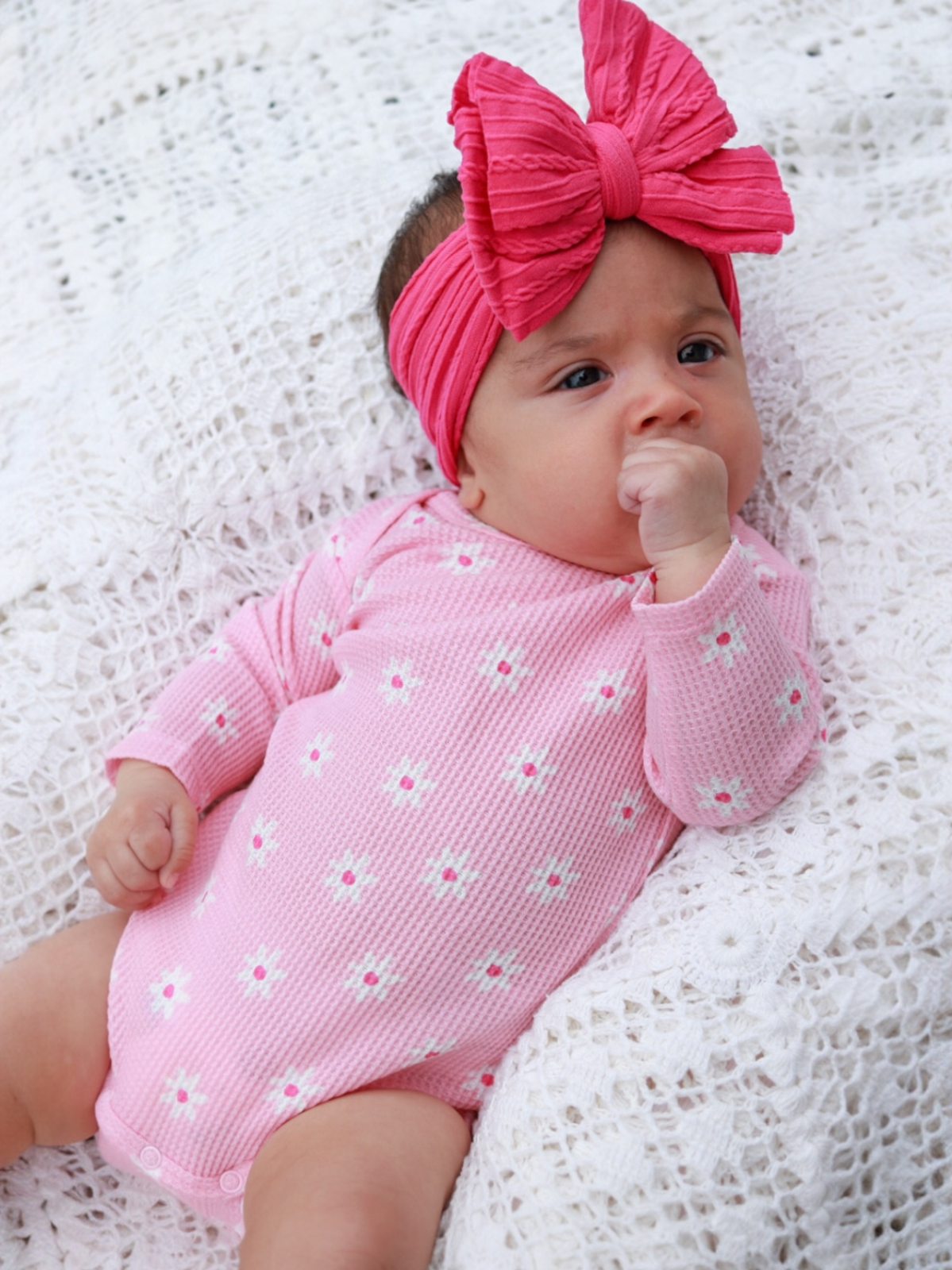 Infant wearing a pink onesie with flowers, sitting on a textured white blanket, with a large pink bow headband.