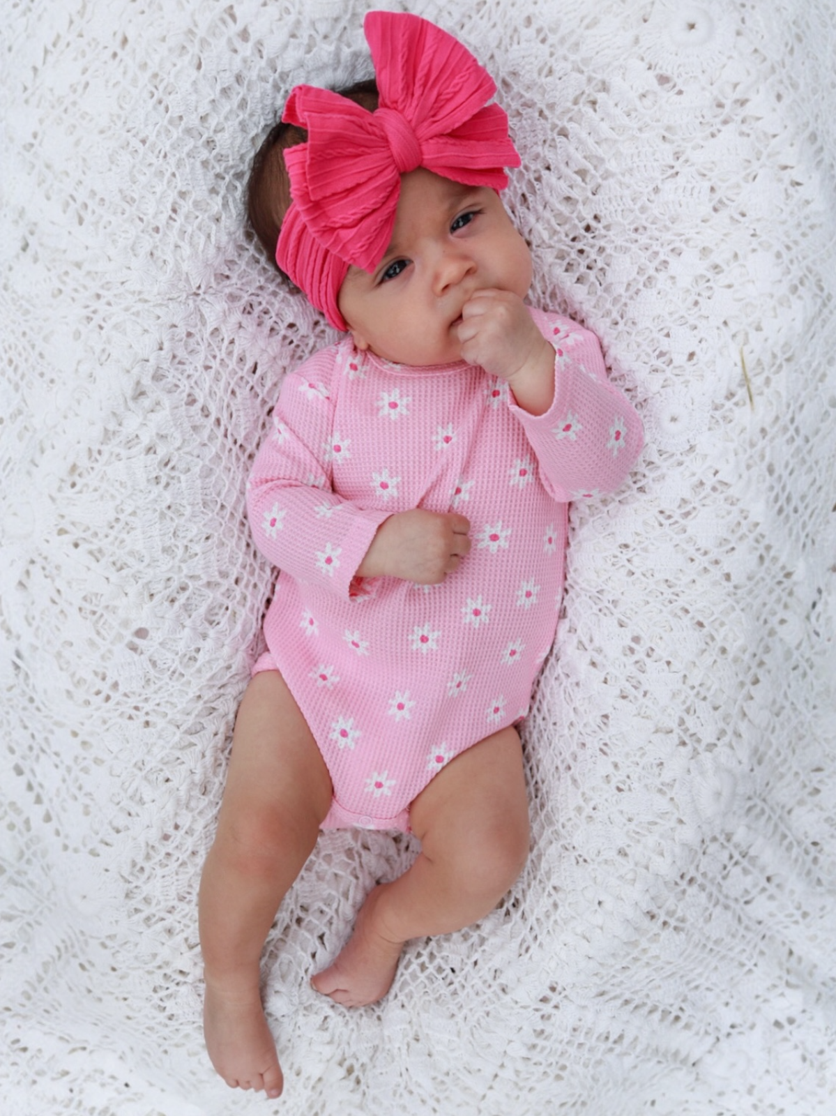 Baby girl in a pink flower-patterned onesie and large pink bow, lying on a textured white blanket.
