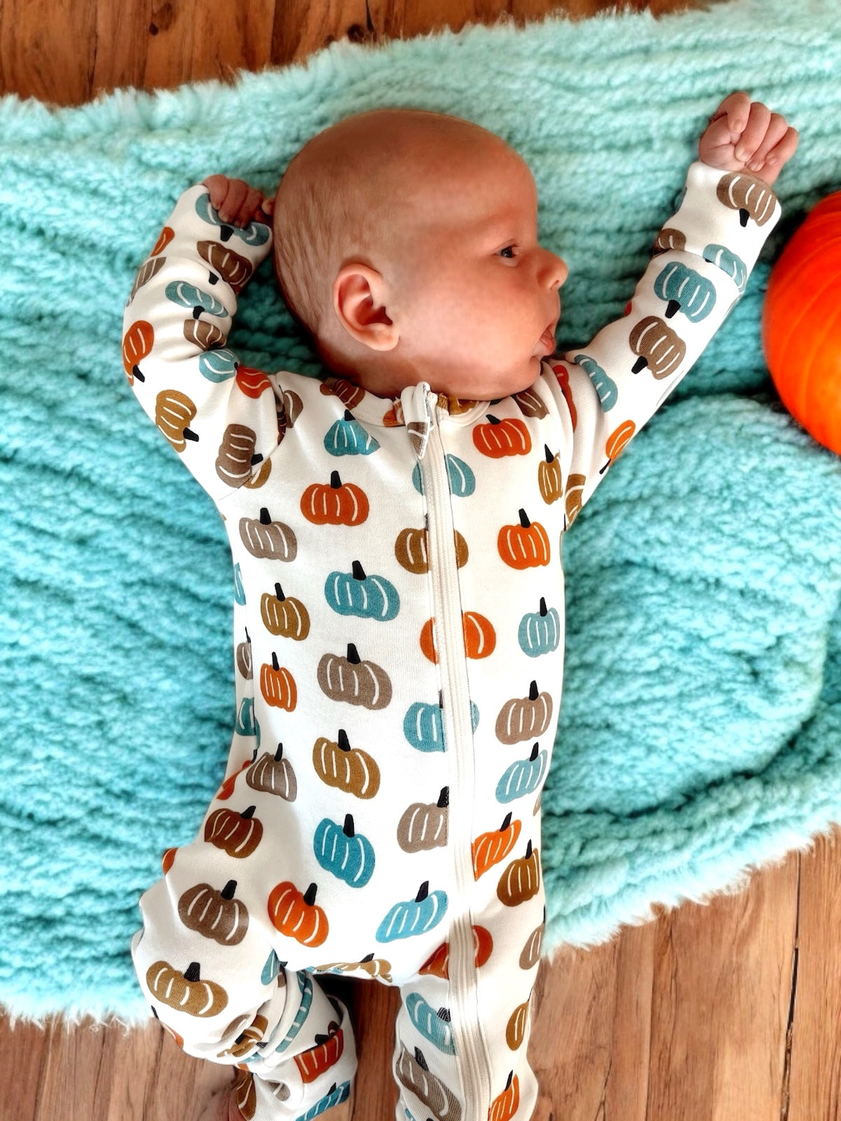 Baby lying on a turquoise blanket, wearing a pumpkin-patterned onesie, with a pumpkin toy nearby.