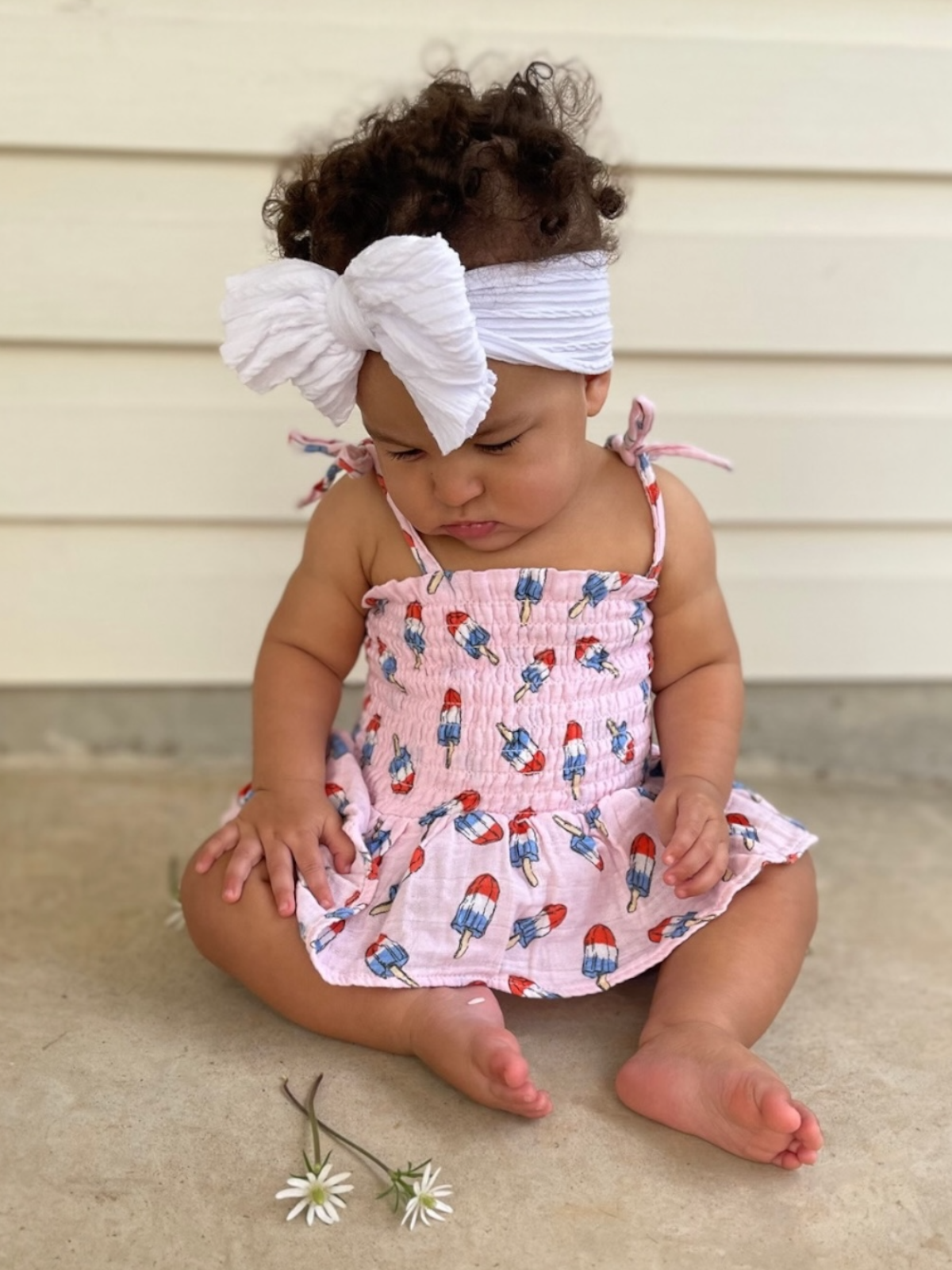 Toddler with curly hair in a pink dress, sitting on the ground, surrounded by daisies.