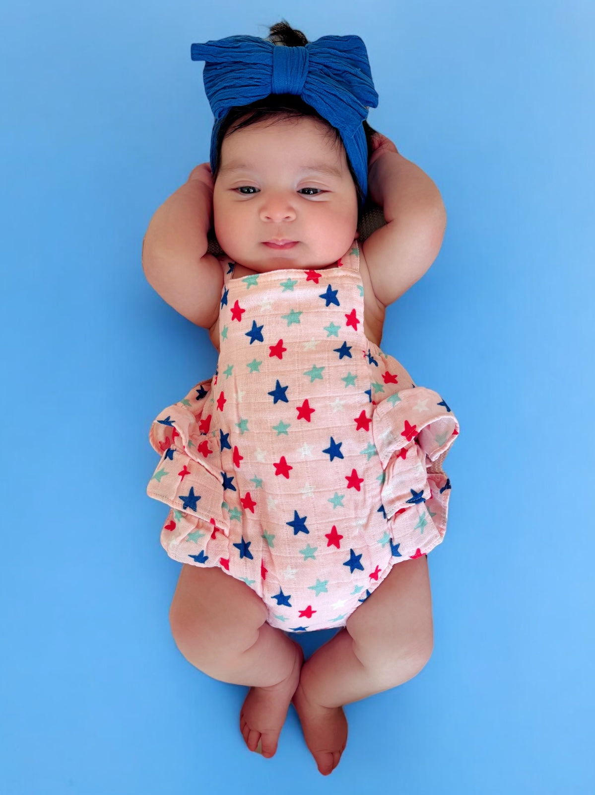 Baby girl in a pastel star-patterned swimsuit and blue bow headband, lying on a blue background.