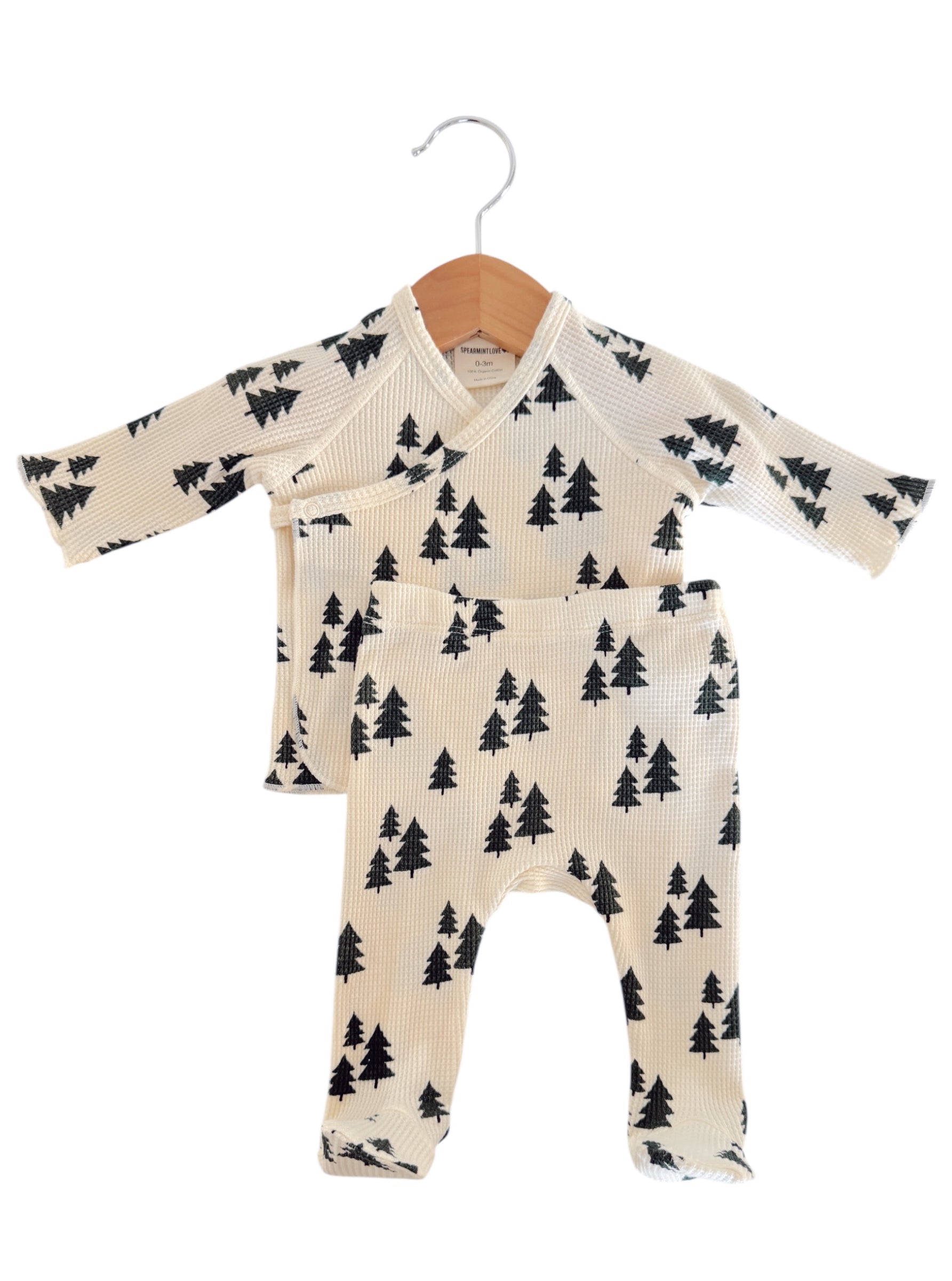 Baby outfit set featuring cream top and pants with black tree print, hanging on a wooden hanger.