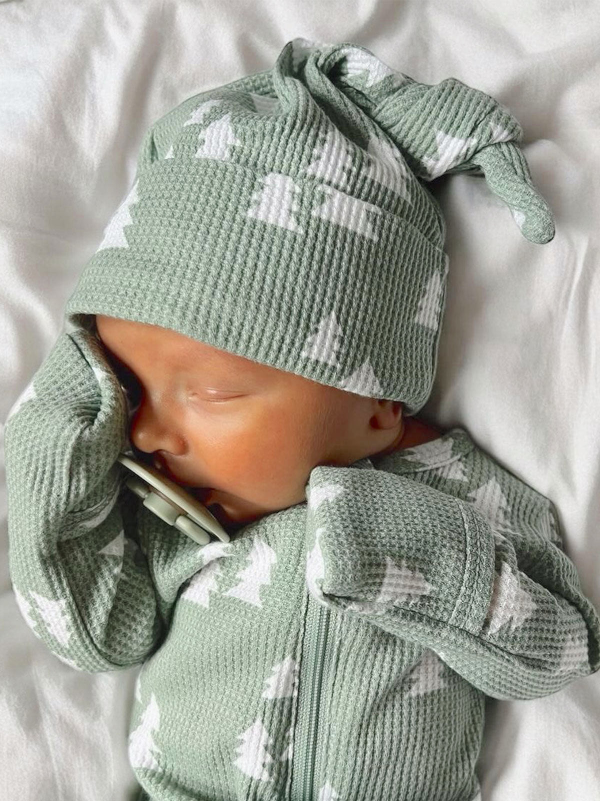 Sleeping baby in a soft green outfit with a knitted hat, holding a pacifier, resting on a white blanket.