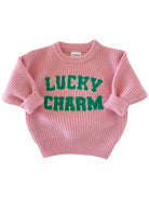 Pink knitted sweater with "LUCKY CHARM" in green text on the front. Cozy and stylish for casual wear.