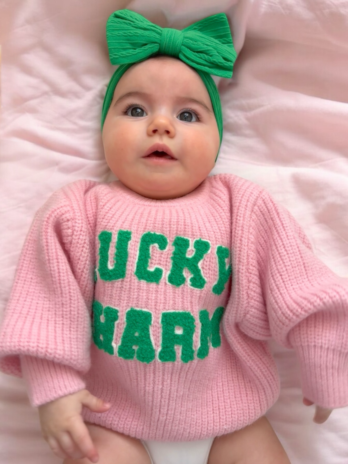 Baby wearing a pink sweater with "LUCKY CHARM" in green, and a green bow headband, lying on a soft pink surface.