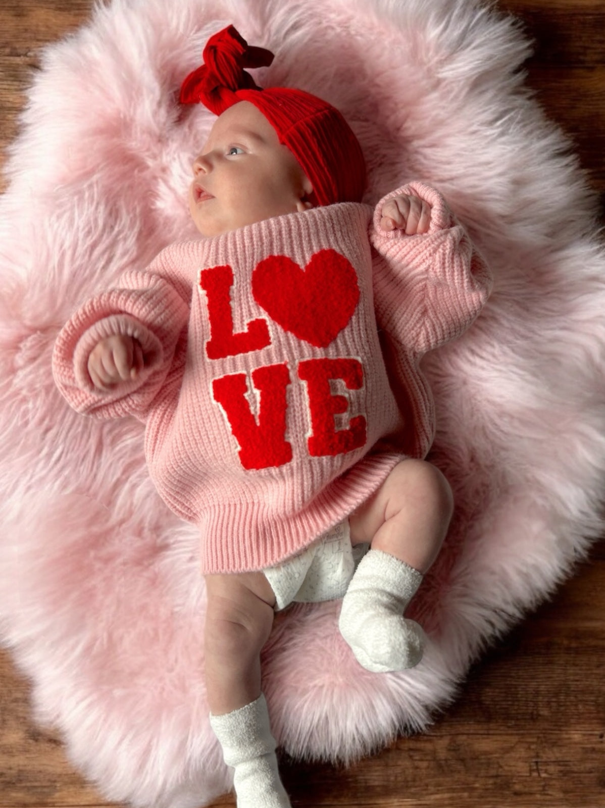 Baby wearing a pink sweater with the text "LOVE" on a fluffy textured rug, with a red headband and white socks.