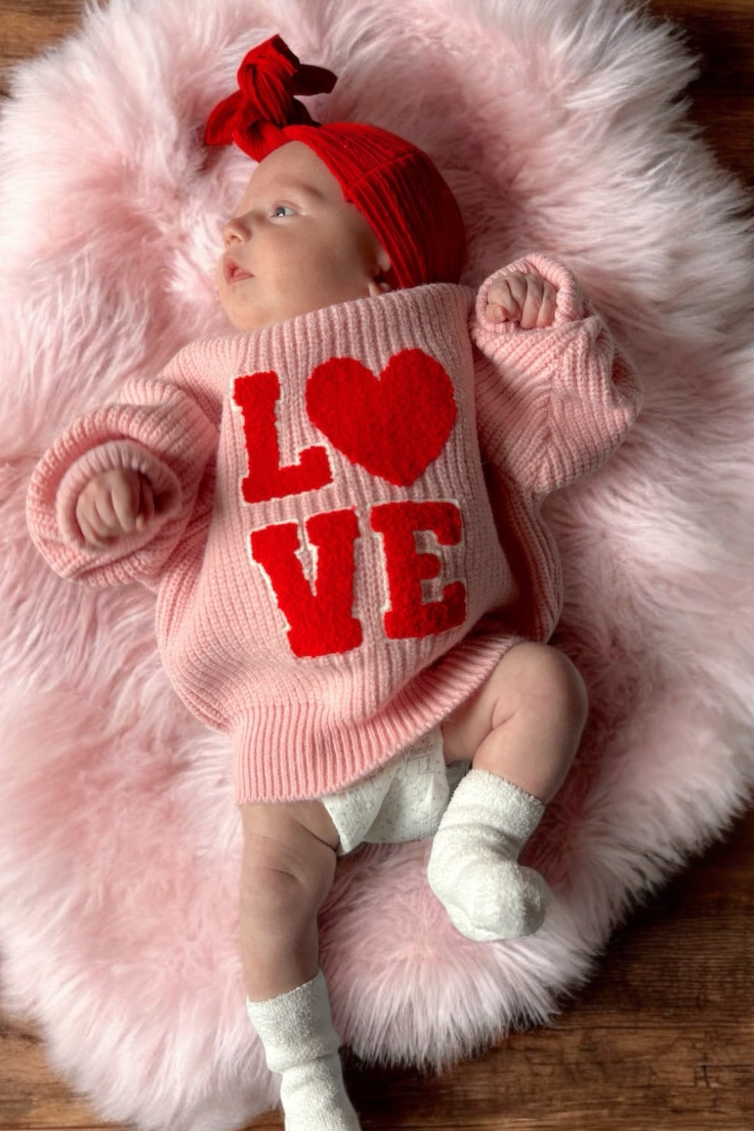 Baby wearing a pink sweater with the text "LOVE" on a fluffy textured rug, with a red headband and white socks.