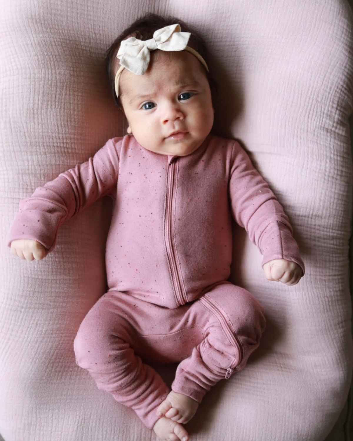 Infant wearing a pink zip-up outfit and bow headband, lying on a soft pink surface, looking curiously at the camera.