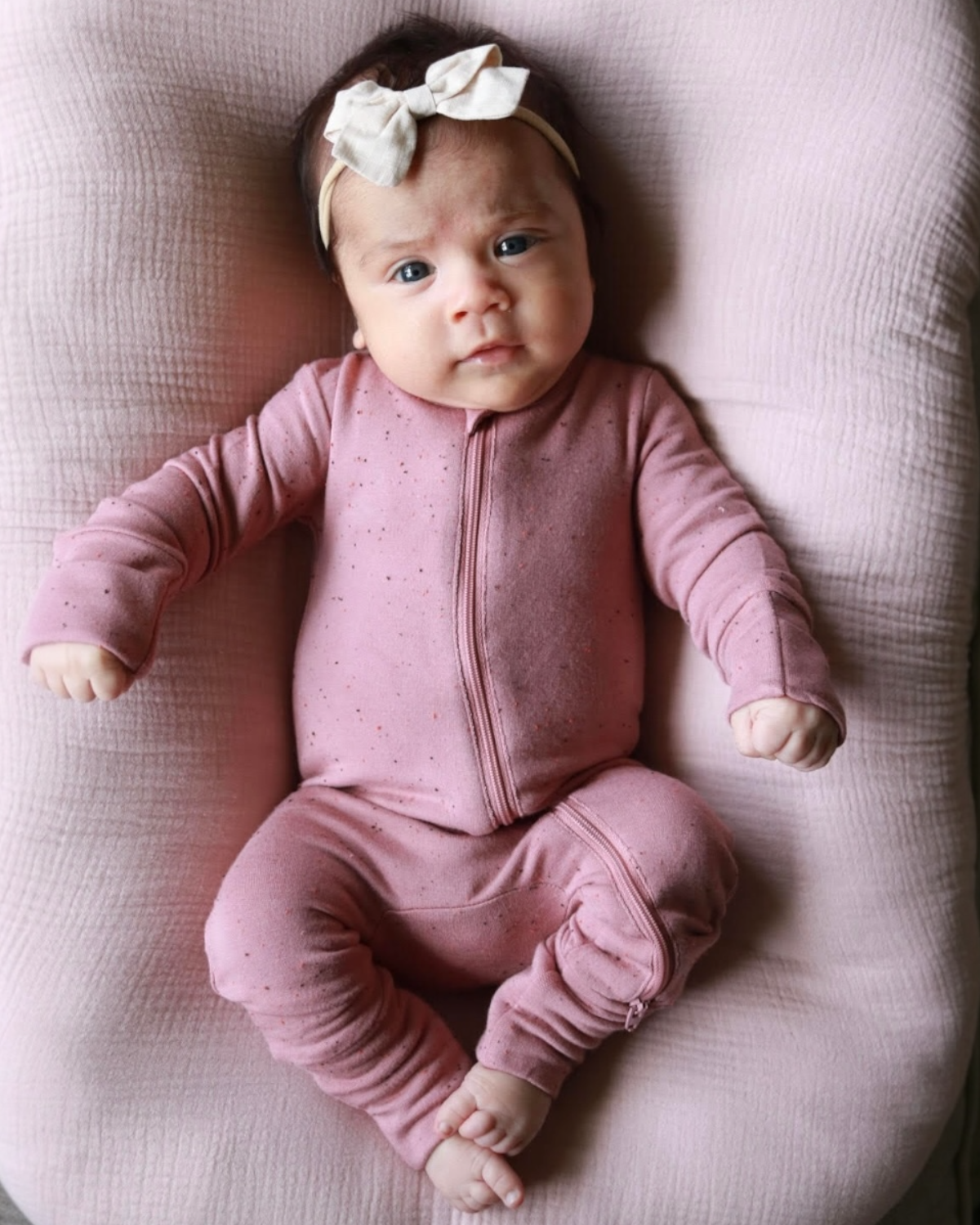 Infant wearing a pink zip-up outfit and bow headband, lying on a soft pink surface, looking curiously at the camera.