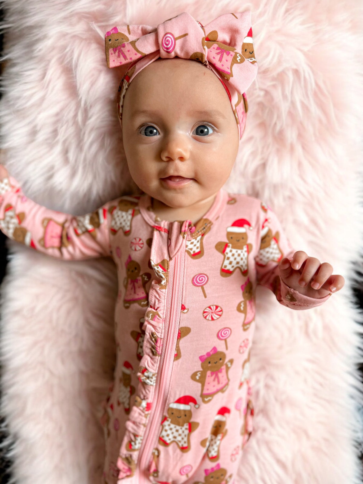 Baby in a pink gingerbread print outfit and bow, lying on a fluffy pink blanket, smiling at the camera.