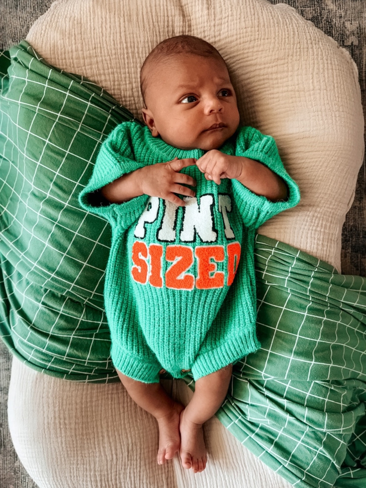 Baby wearing a green "Pint Sized" outfit, lying on a soft surface with green patterned pillows around.