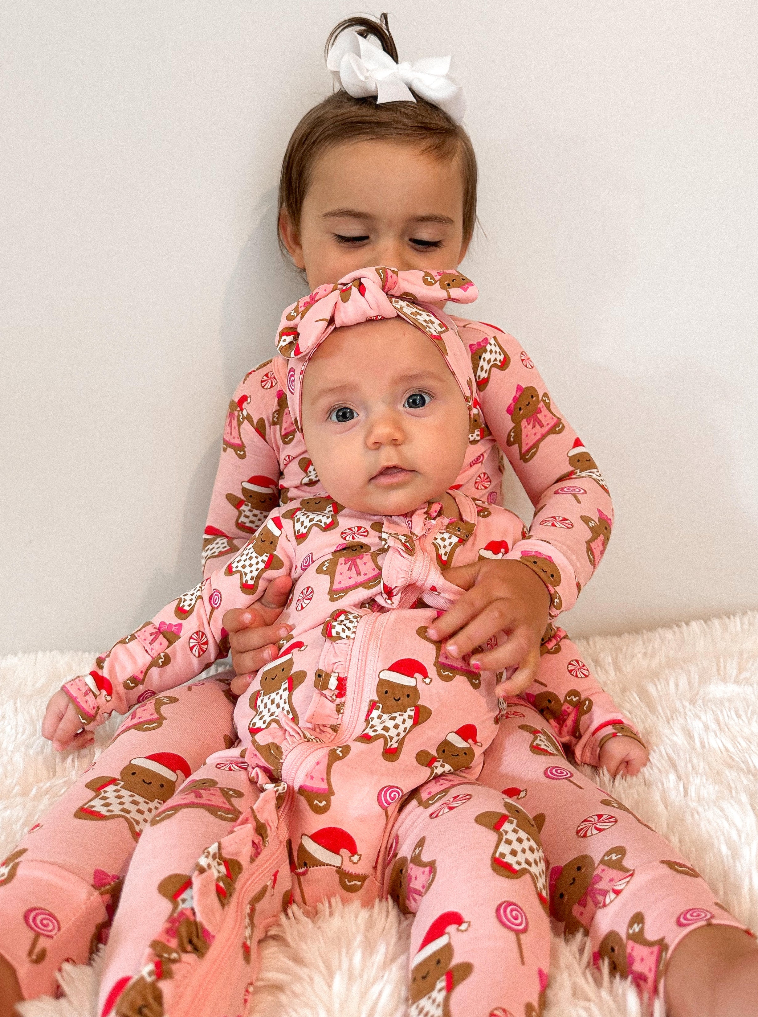 Two young siblings in matching pink gingerbread-themed pajamas, sitting on a soft white surface.