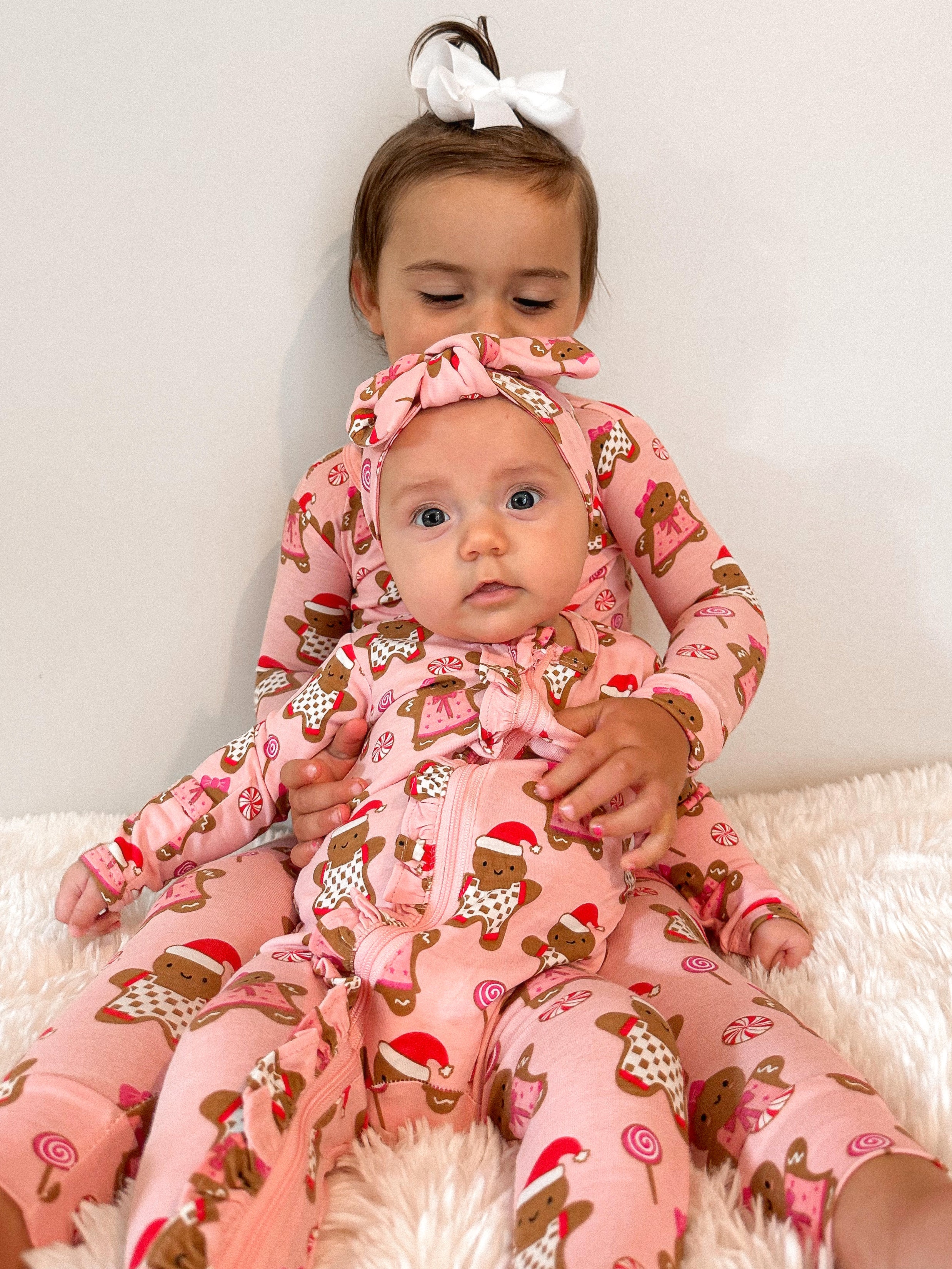 Two young siblings in matching pink gingerbread-themed pajamas, sitting on a soft white surface.