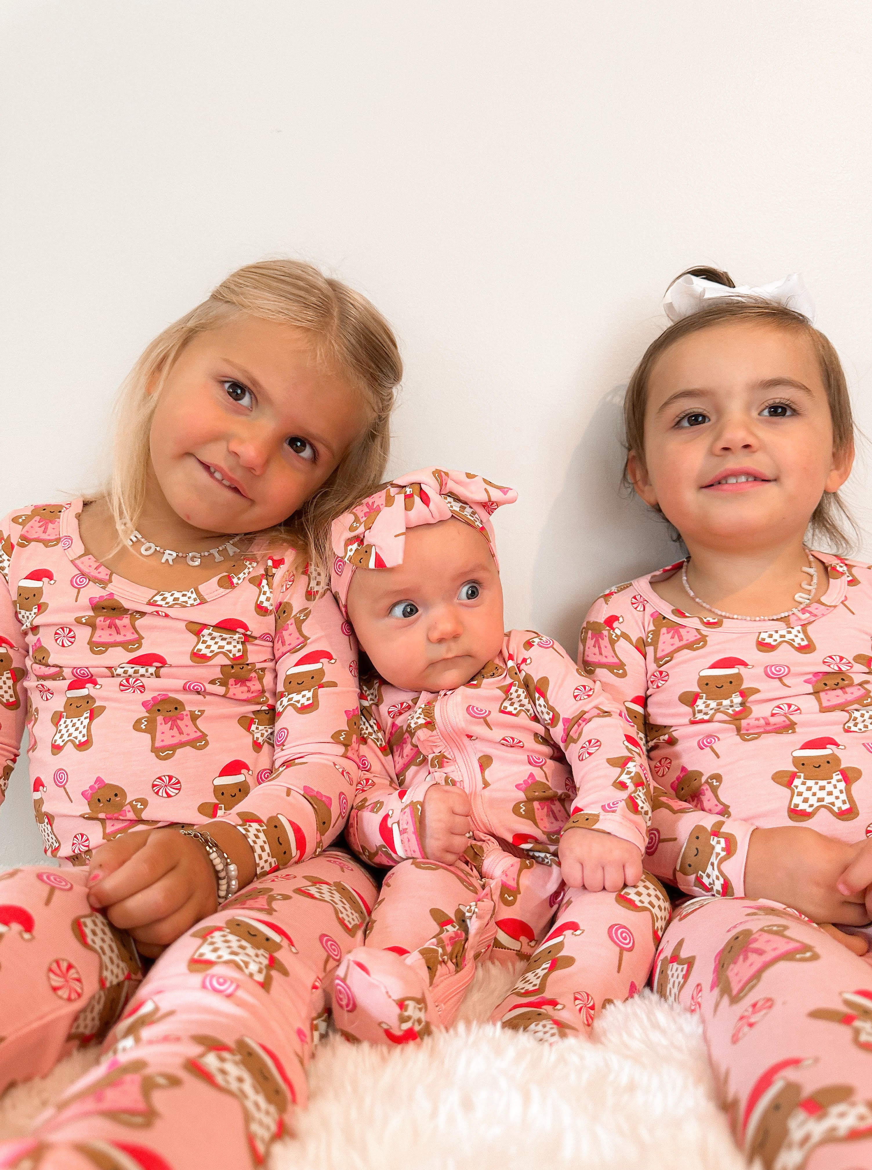 Three young girls in festive pink pajamas with gingerbread motifs, sitting together against a white background.
