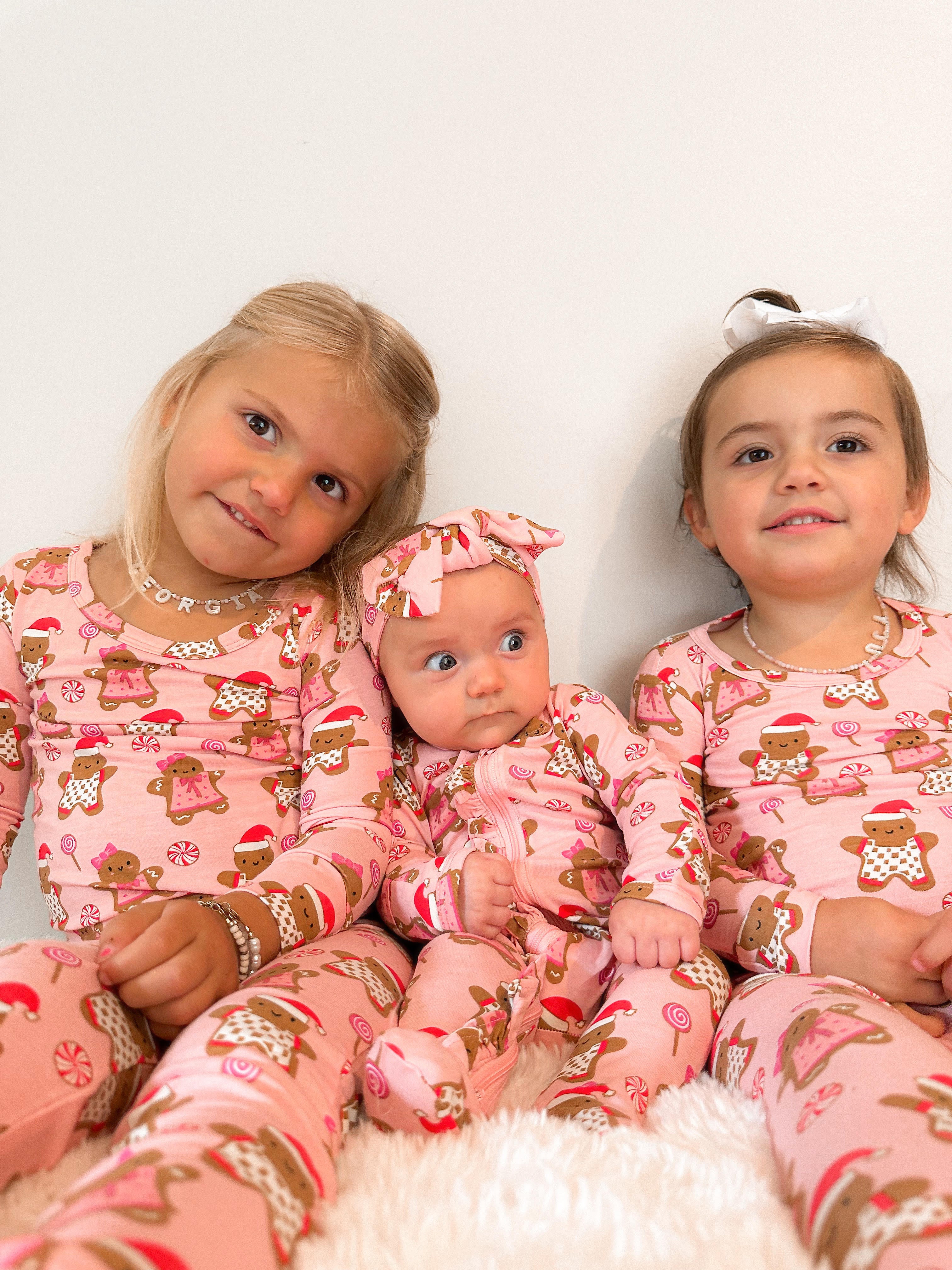 Three young girls in festive pink pajamas with gingerbread motifs, sitting together against a white background.