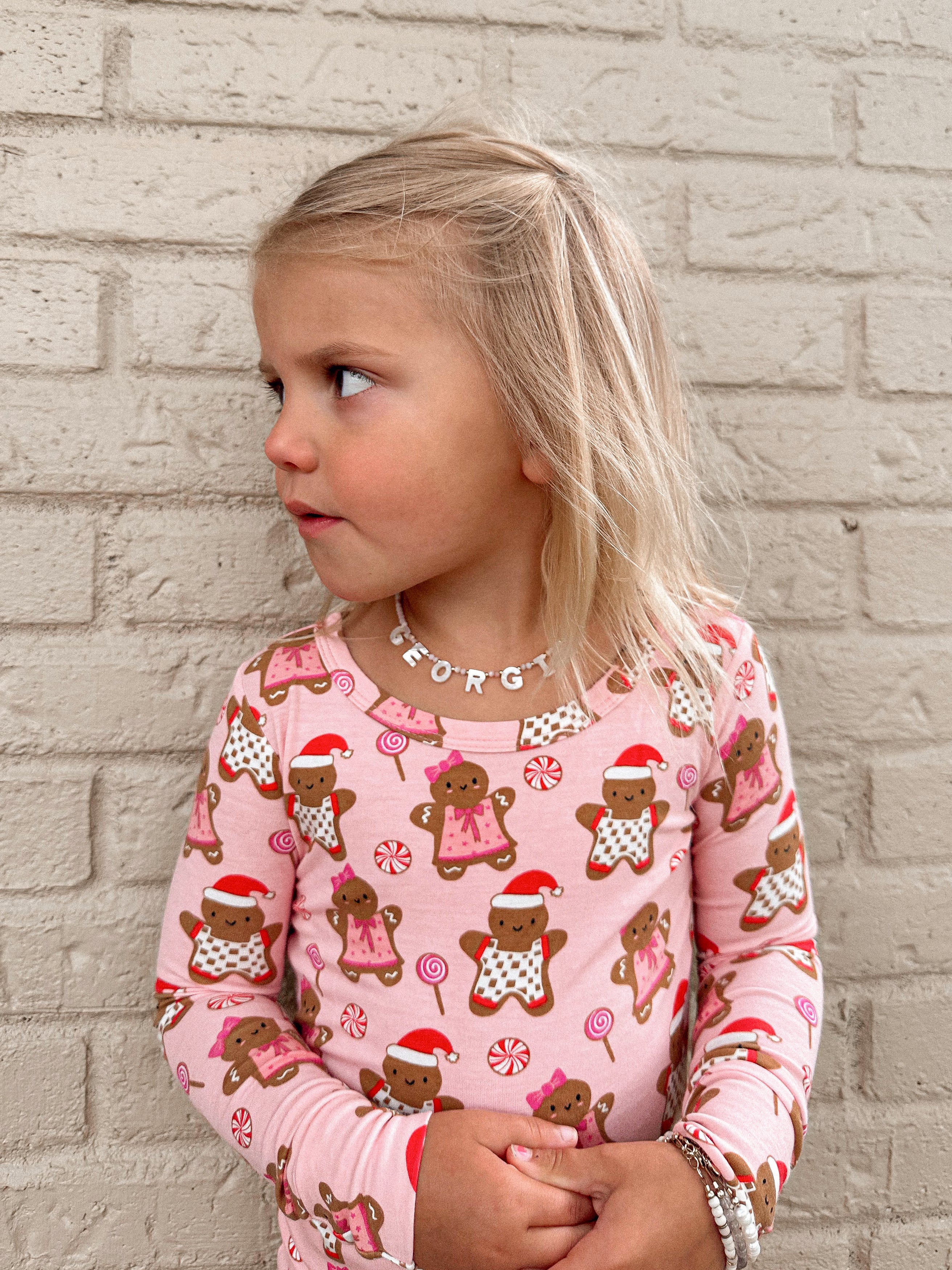 Young girl in a pink gingerbread-patterned shirt, looking sideways against a brick wall background.