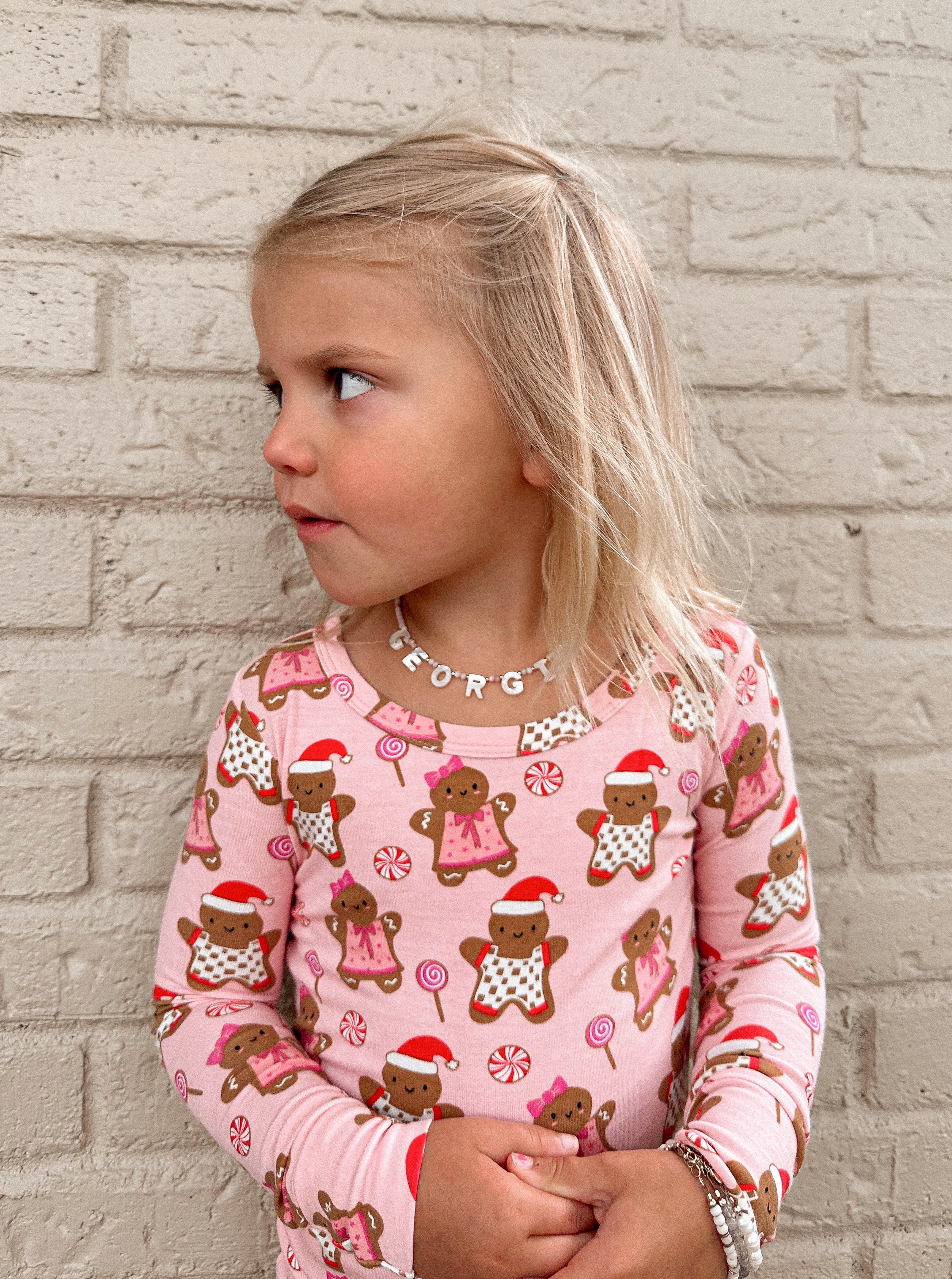 Young girl in a pink gingerbread-patterned shirt, looking sideways against a brick wall background.