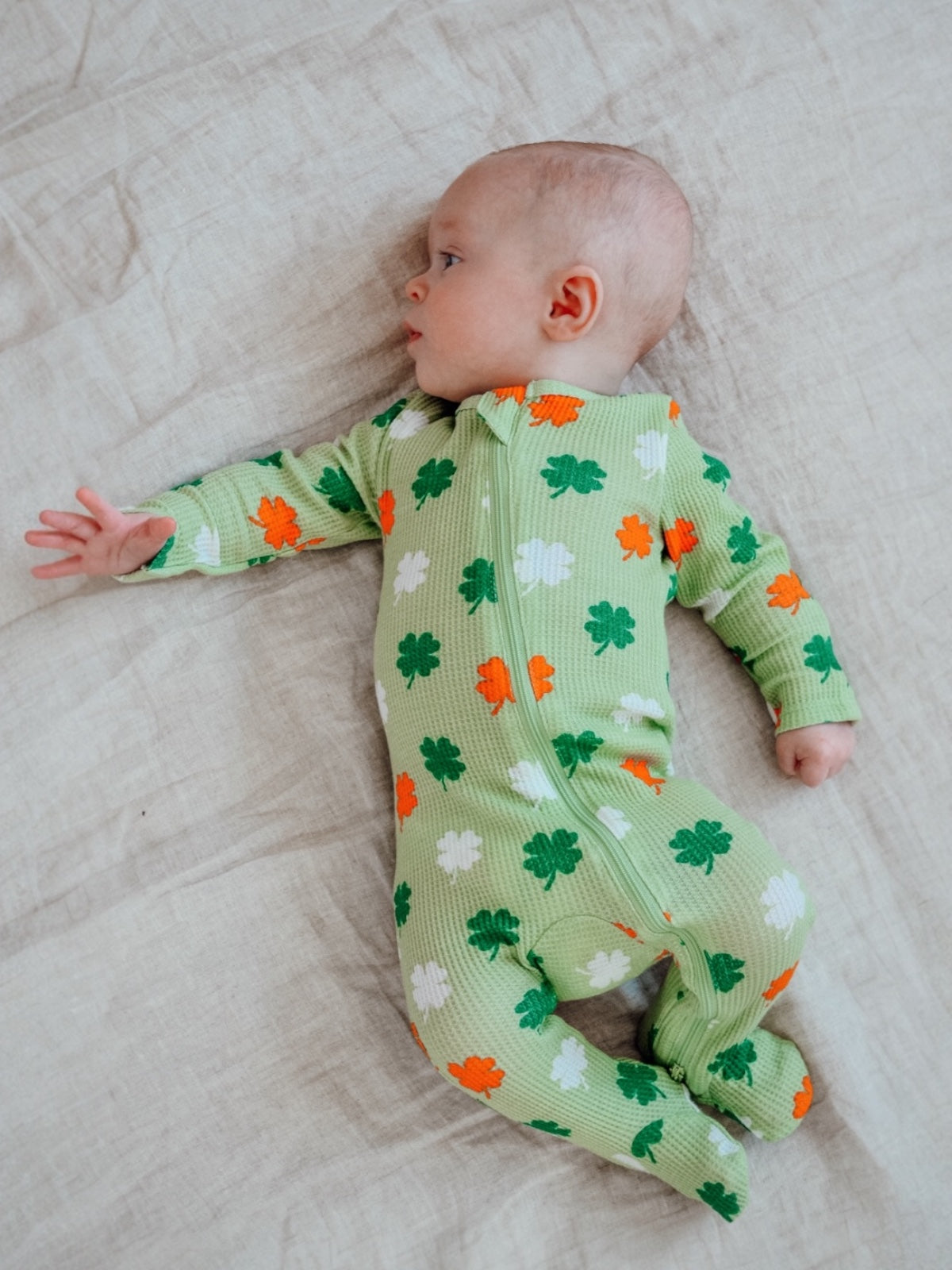 Baby in green shamrock-patterned onesie lying on light-colored fabric.