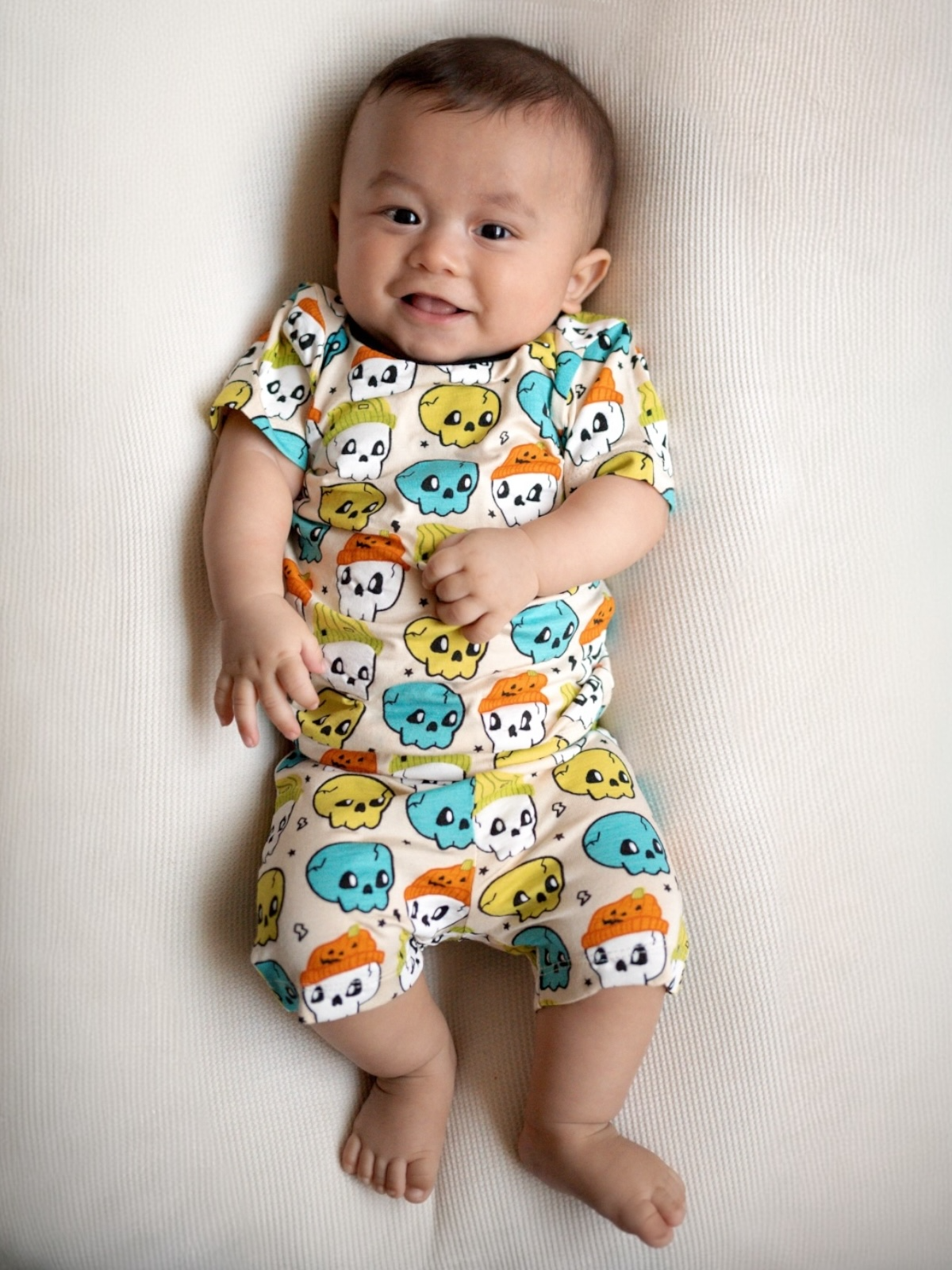 Smiling baby in colorful skull-patterned outfit lying on a soft surface.