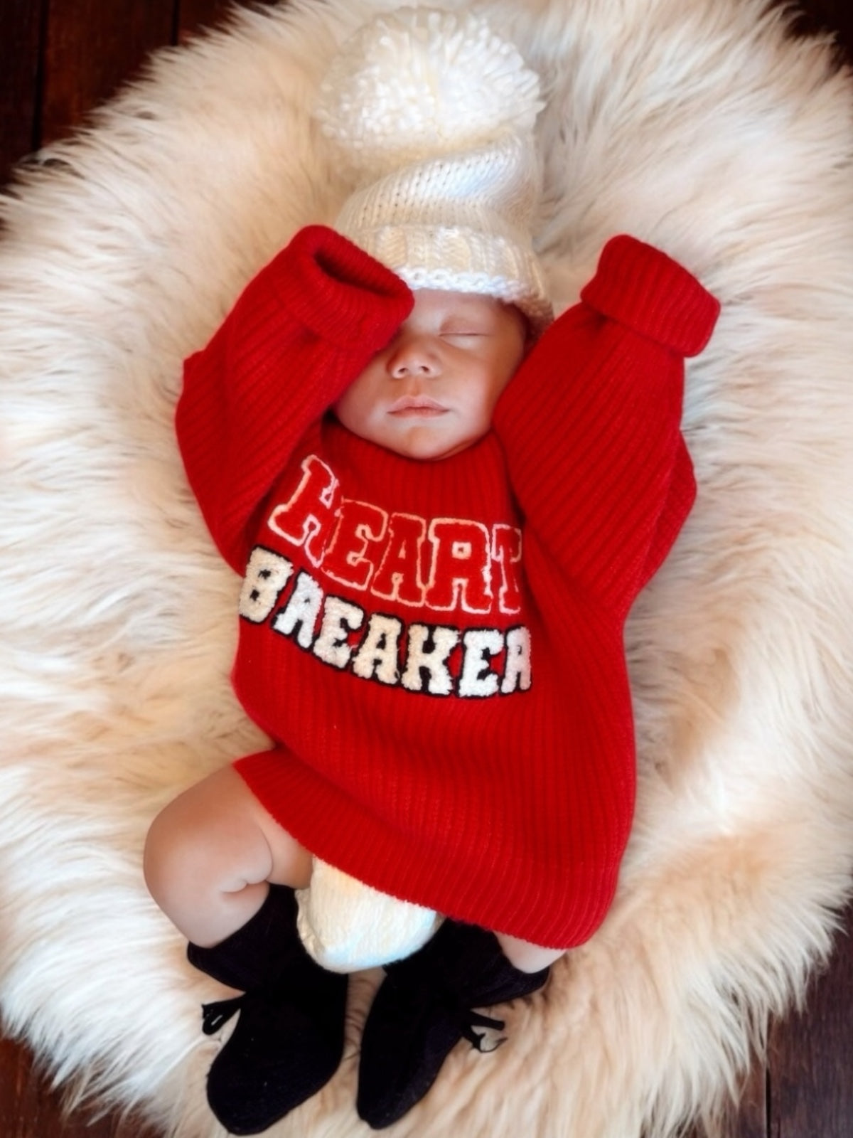 Baby in a red "Heartbreaker" sweater and white hat, lying on a fluffy rug, hands raised, looking peaceful.