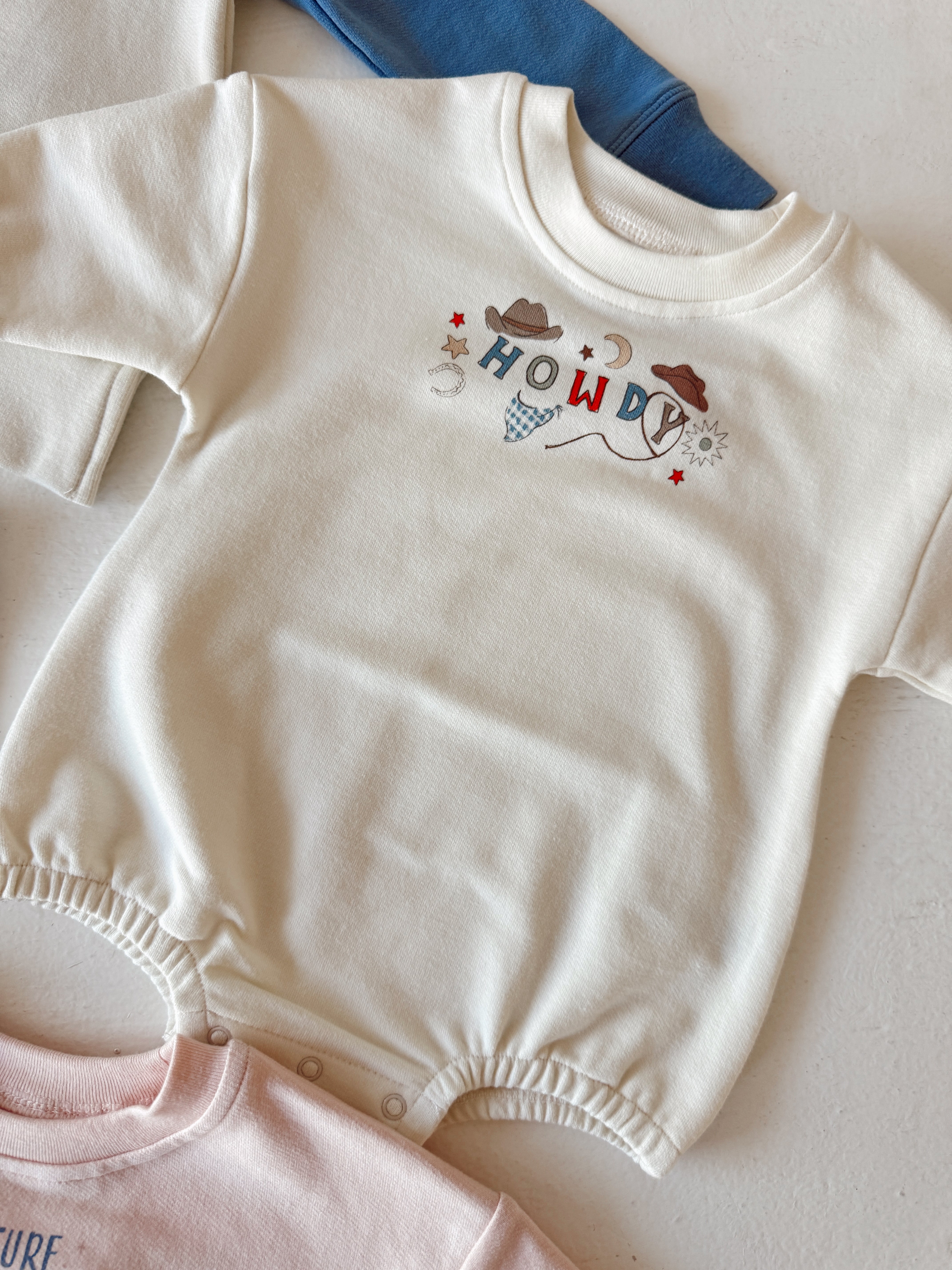 Cream-colored baby onesie with "HOWDY!" design and decorative elements, laid out with other colorful shirts.