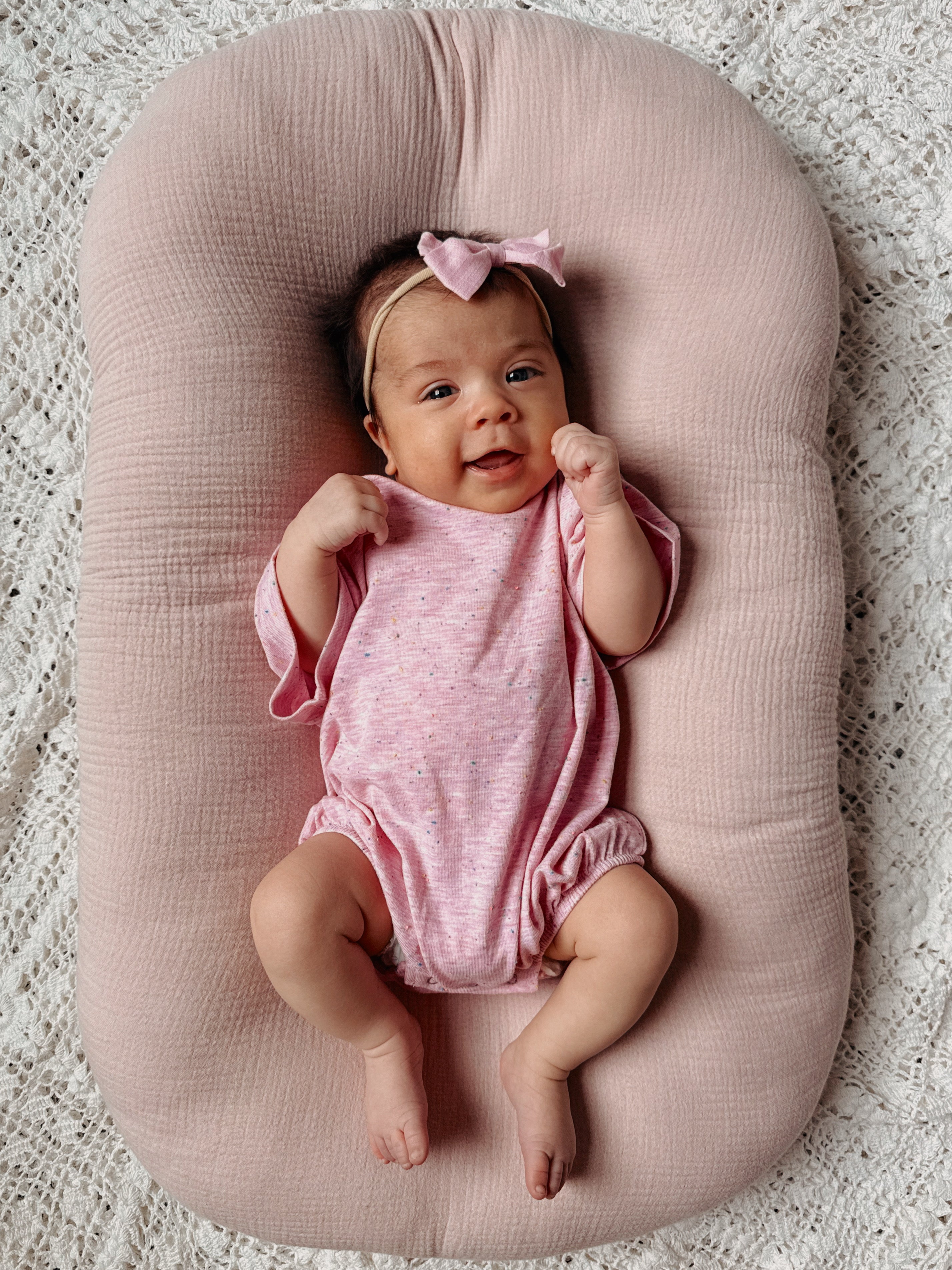 Smiling baby girl in a pink onesie with a bow headband, nestled in a soft pink cushion on a lace blanket.