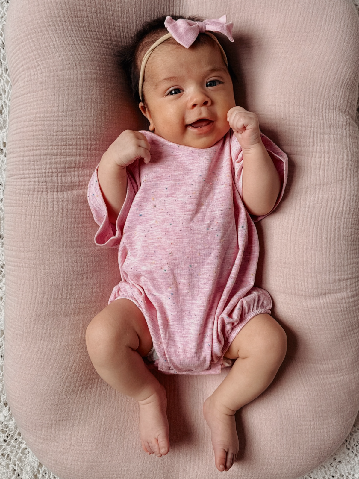 Smiling baby girl in pink onesie with bow, resting on soft pink support pillow.