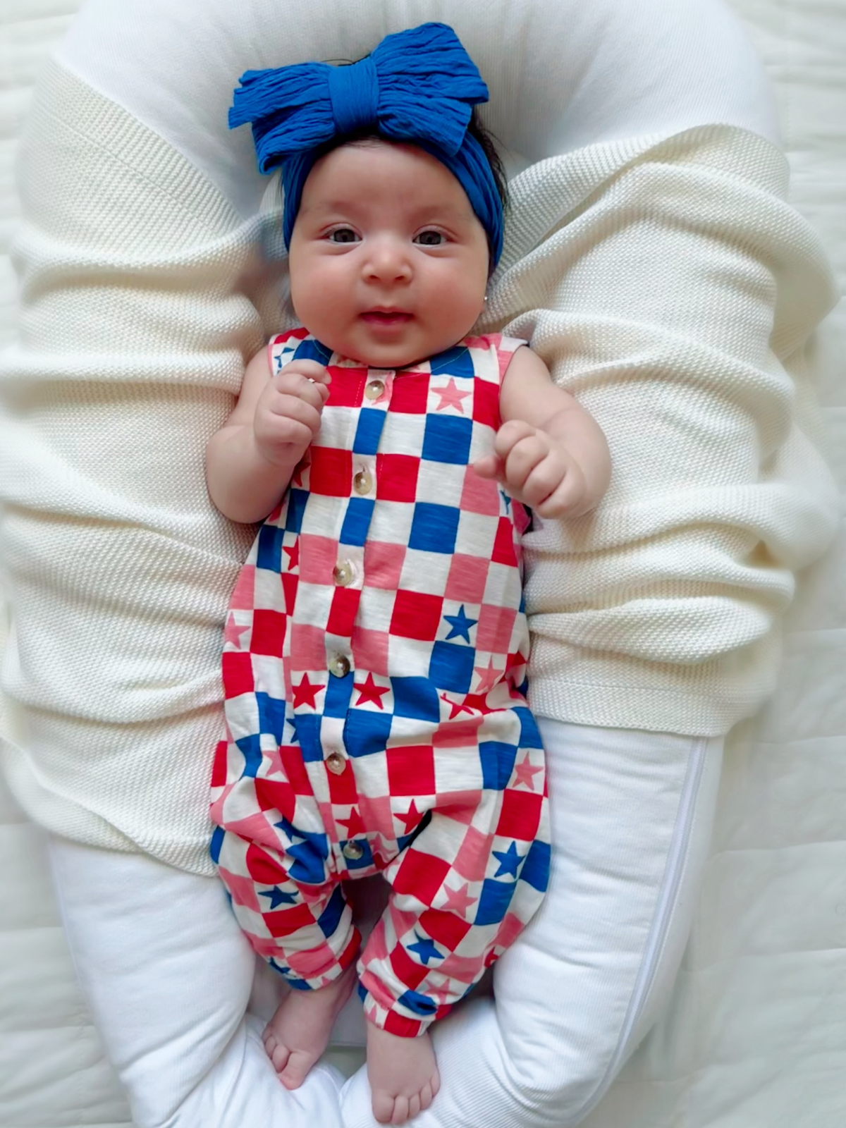 Baby in a red, white, and blue star-patterned outfit with a blue headband, smiling on a cozy blanket.