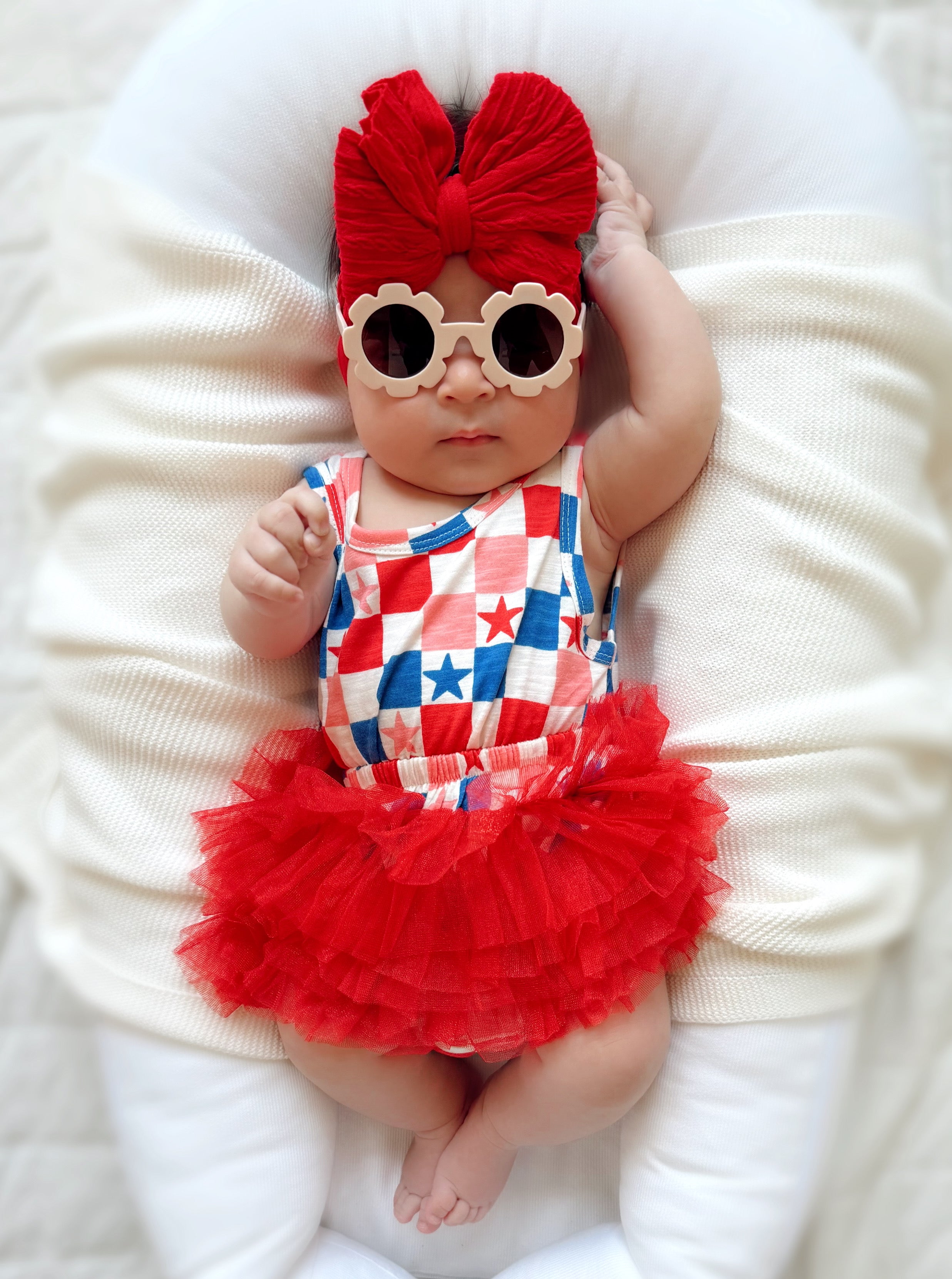 Baby in a red tutu and star-patterned outfit, wearing oversized sunglasses and a large red bow, relaxing on a soft surface.
