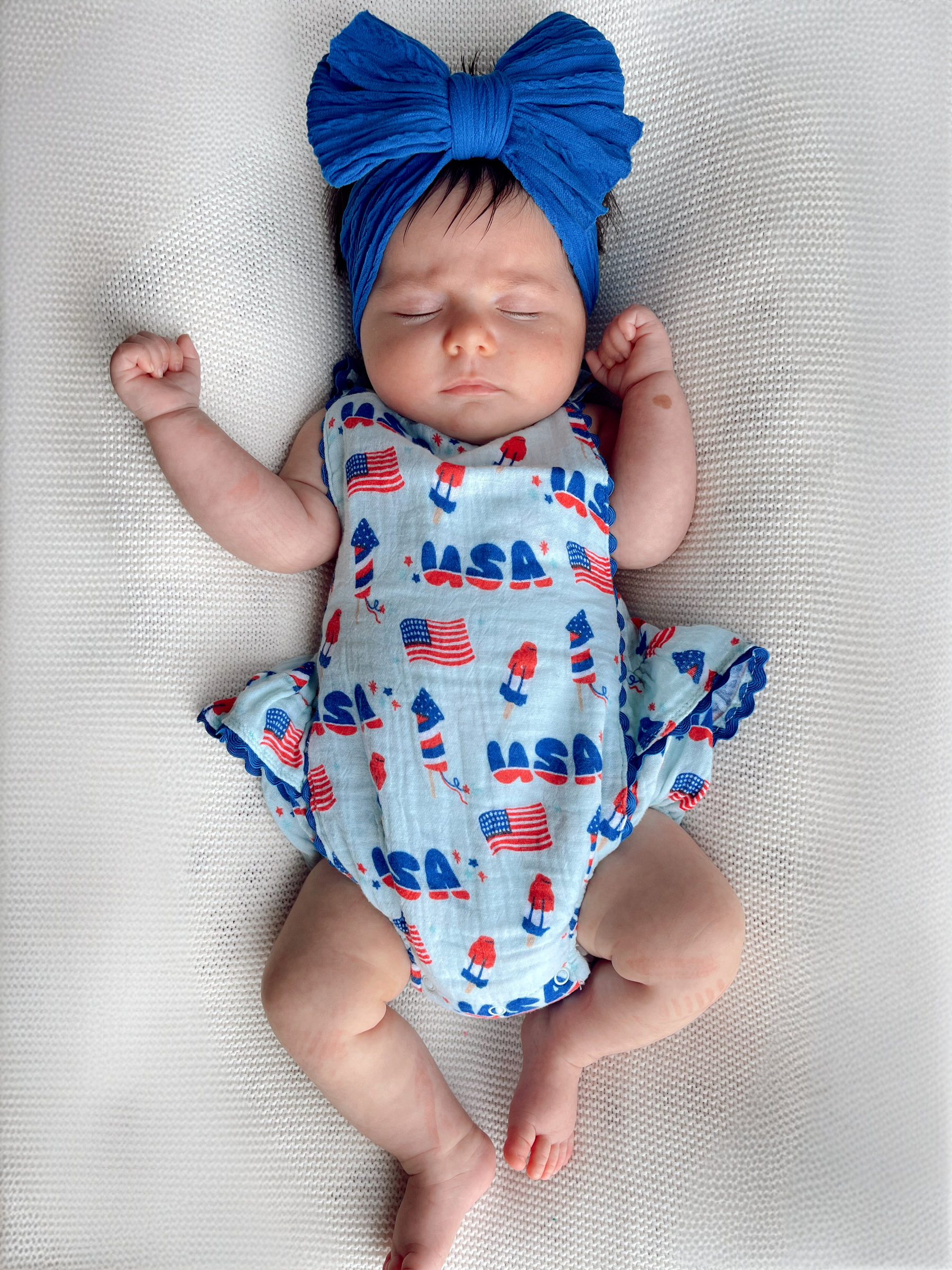 Sleeping baby in a blue headband and patriotic outfit, lying on a textured white surface.