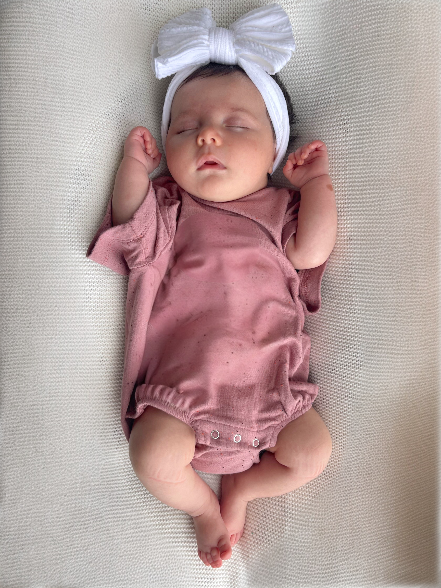 Baby girl in a pink onesie and white headband, sleeping peacefully on a soft, textured blanket.