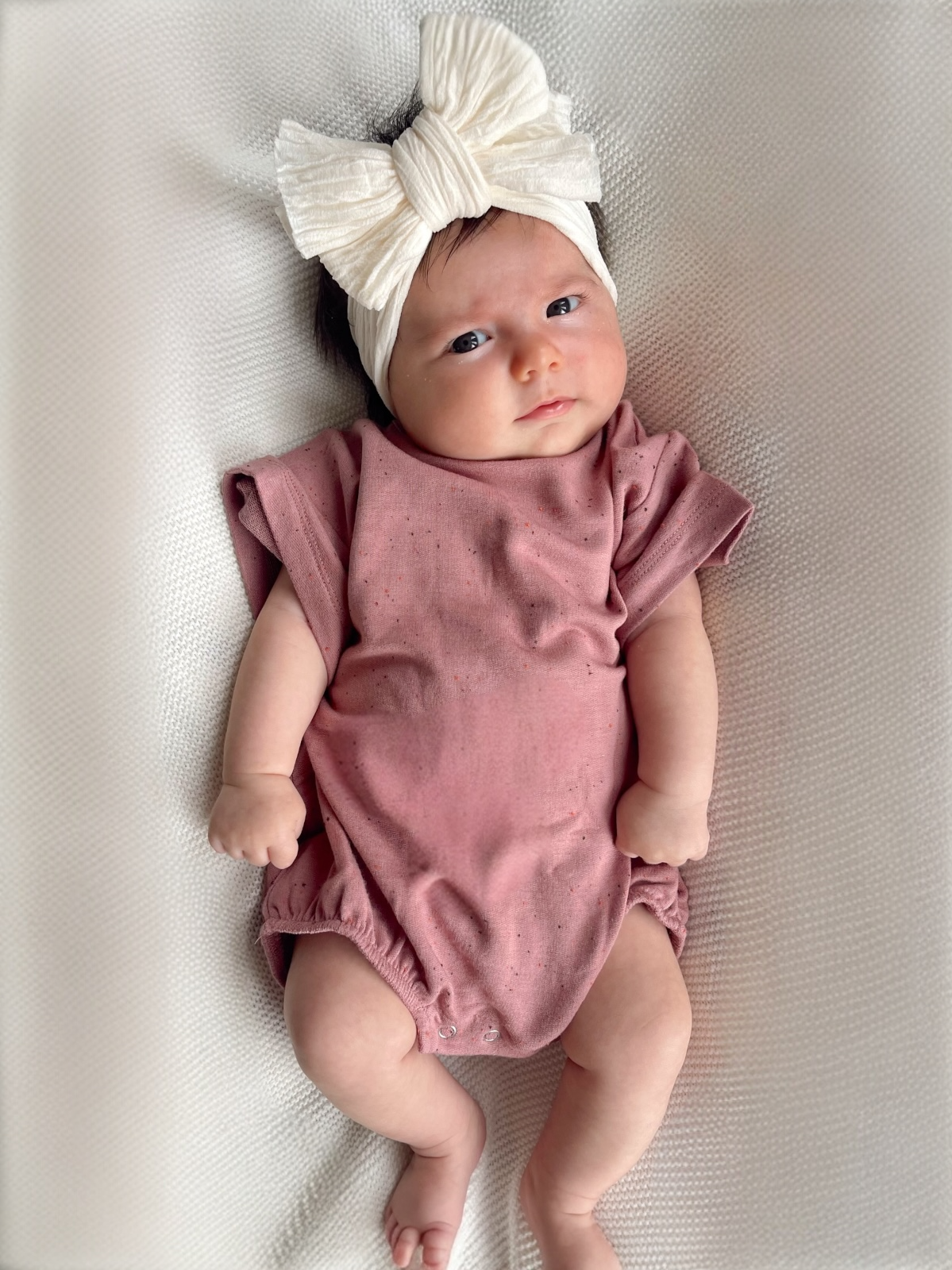 Infant girl in a pink onesie, with a large white bow headband, lying on a textured light background.