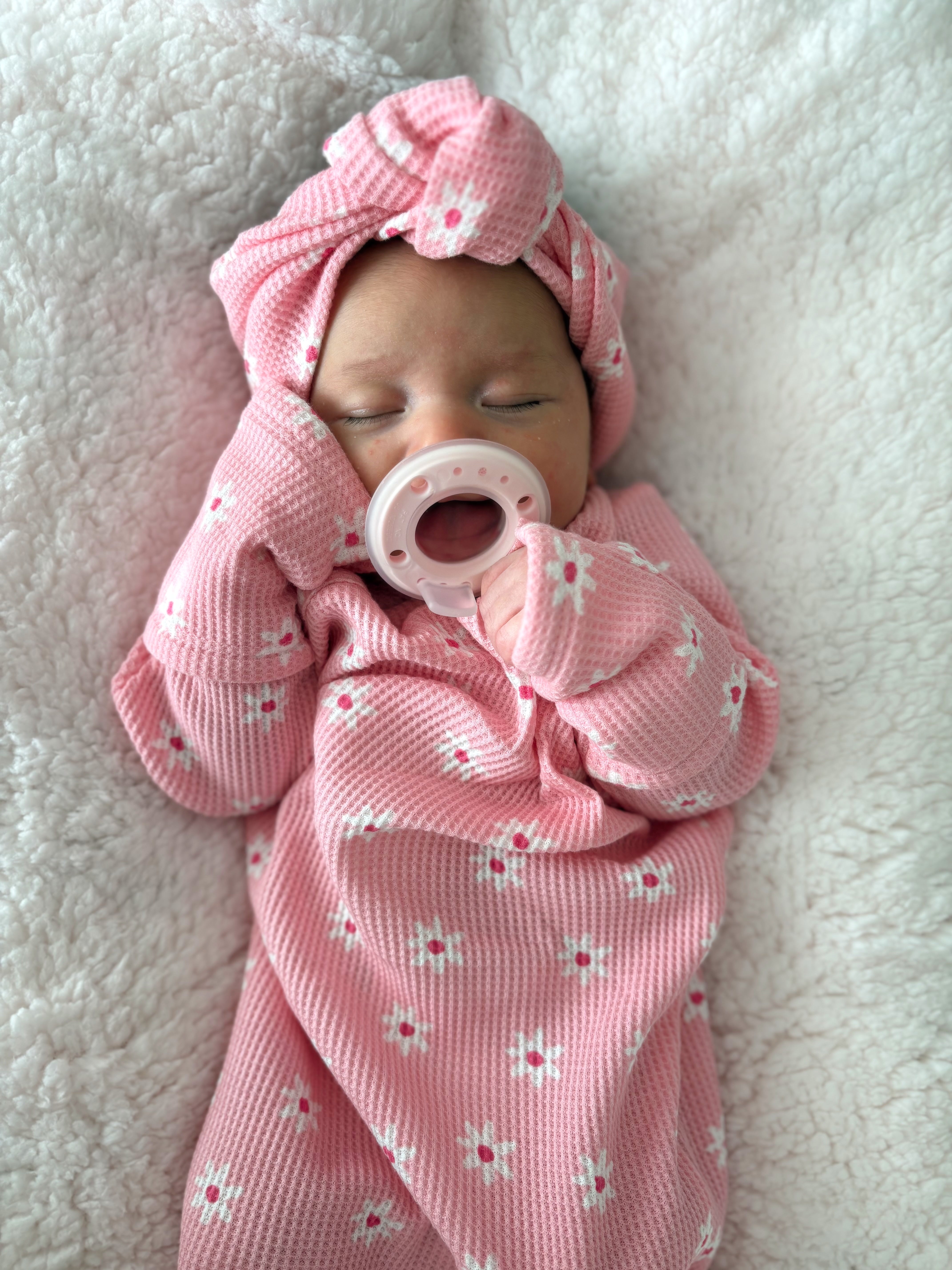 Newborn baby girl in a pink floral outfit and hat with a pacifier, resting on a soft white blanket.