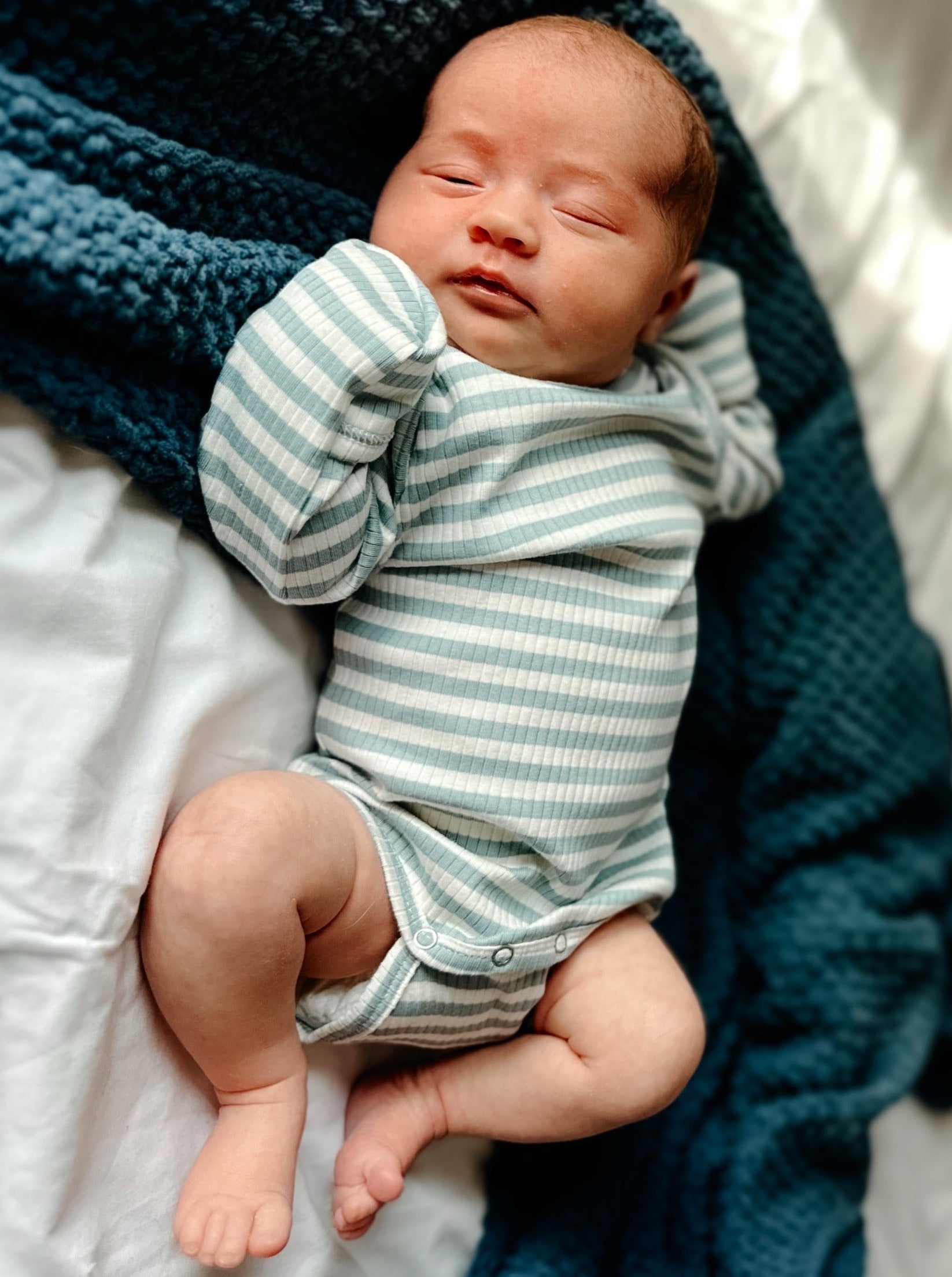 Newborn baby in striped onesie resting on a blue blanket, hands stretched out, relaxed expression.