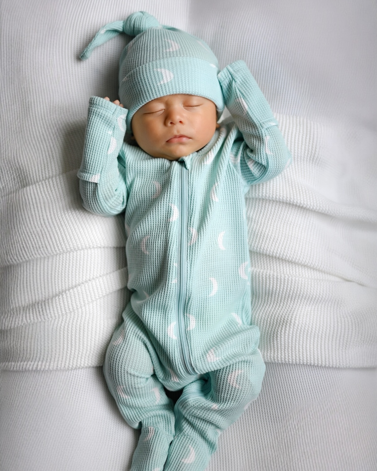 Sleeping baby in mint green pajamas with moon patterns, lying on a textured white blanket.