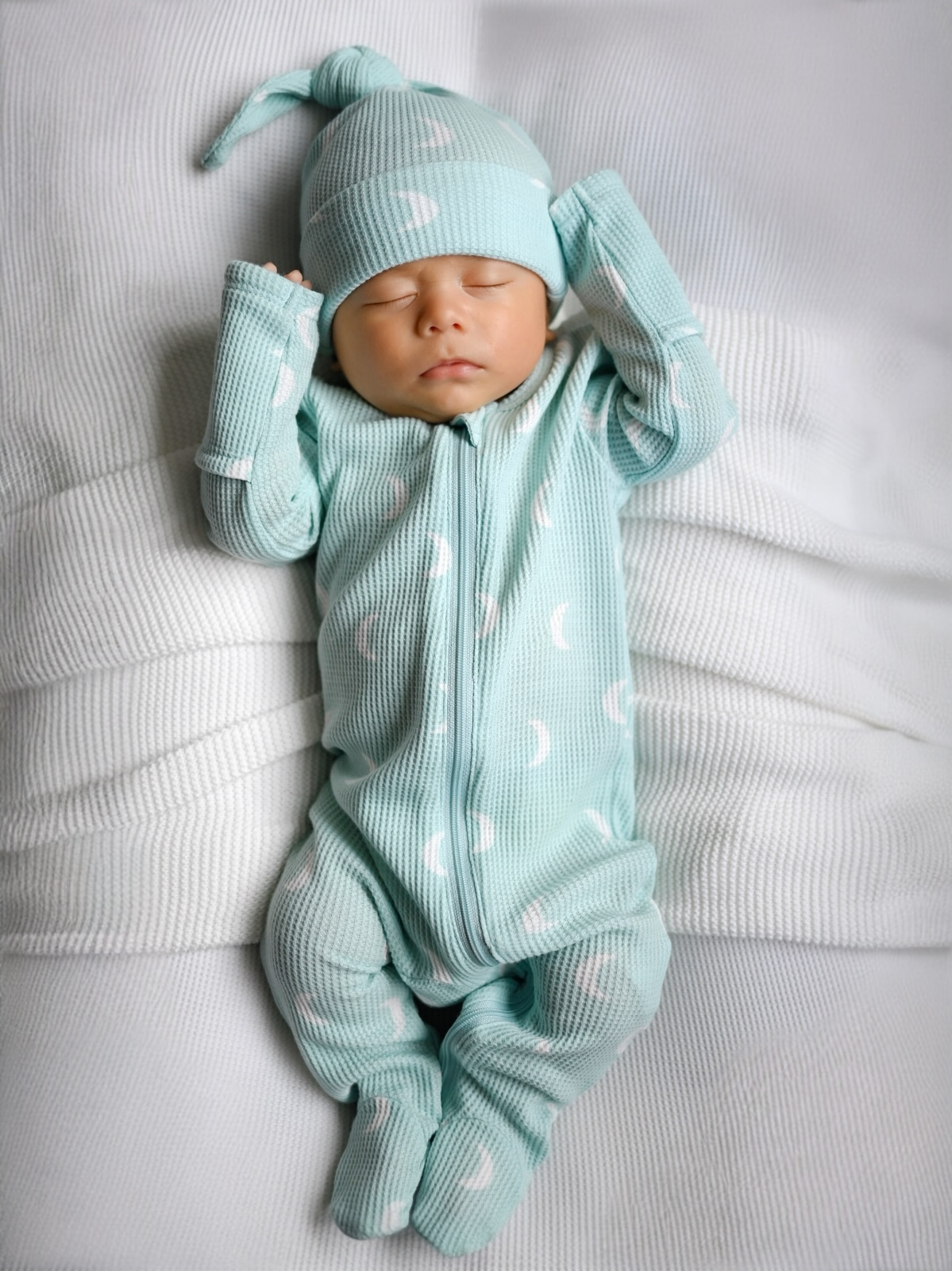 Sleeping baby in mint green pajamas with moon patterns, lying on a textured white blanket.