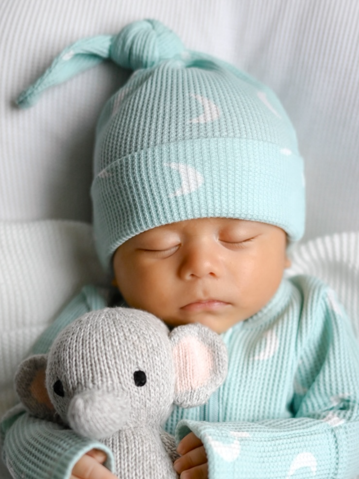 Sleeping baby in a mint green knit hat, holding a gray elephant toy, on a soft white blanket.