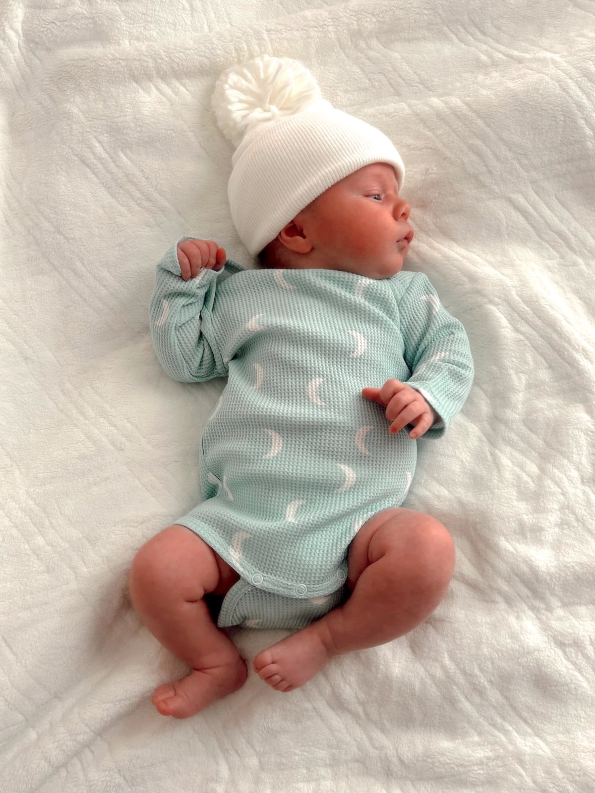 Newborn in a mint green outfit and white beanie, lying on a soft, textured blanket.